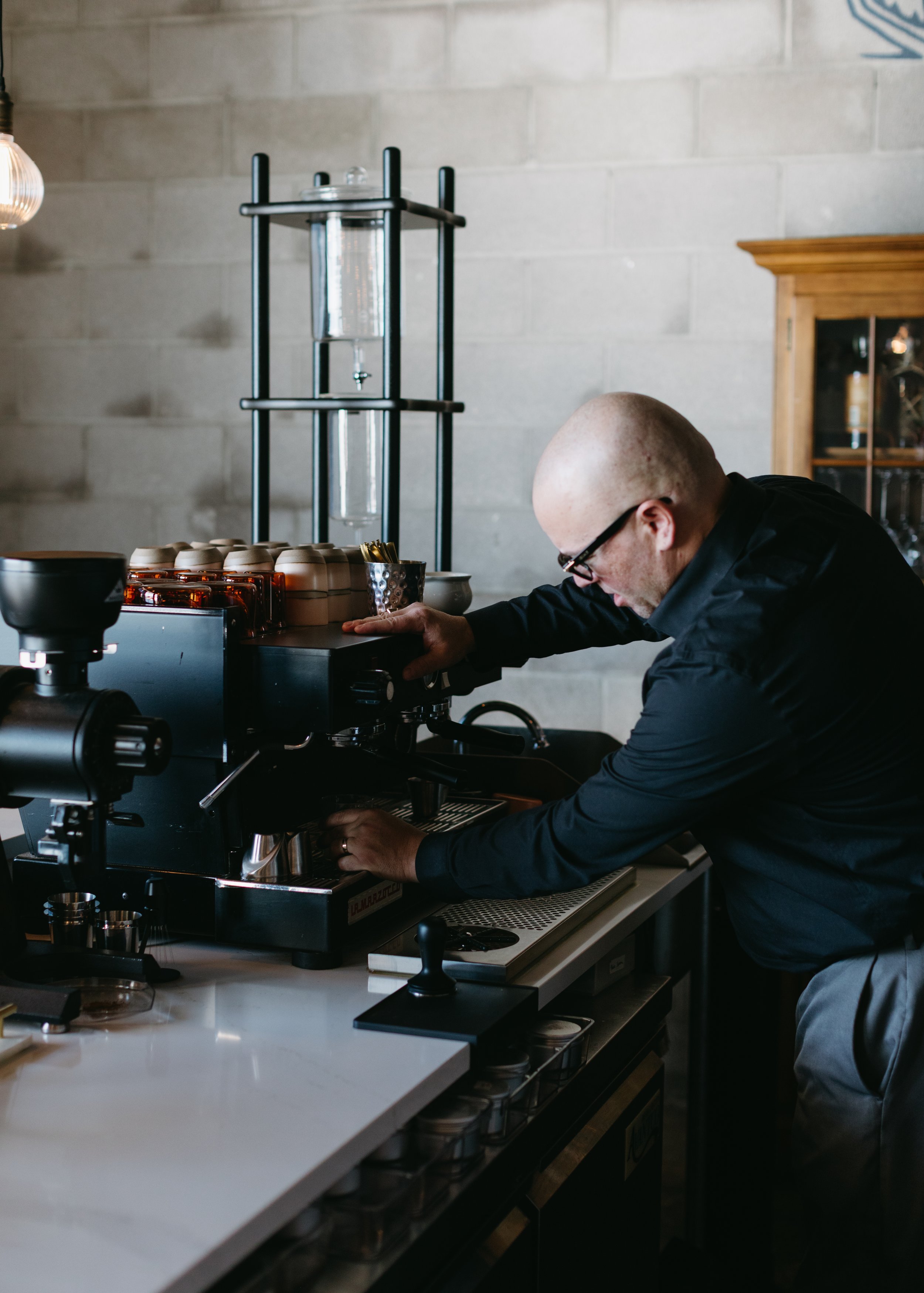 A bald man wearing glasses and a black shirt making espresso at a coffee shop with modern decor.