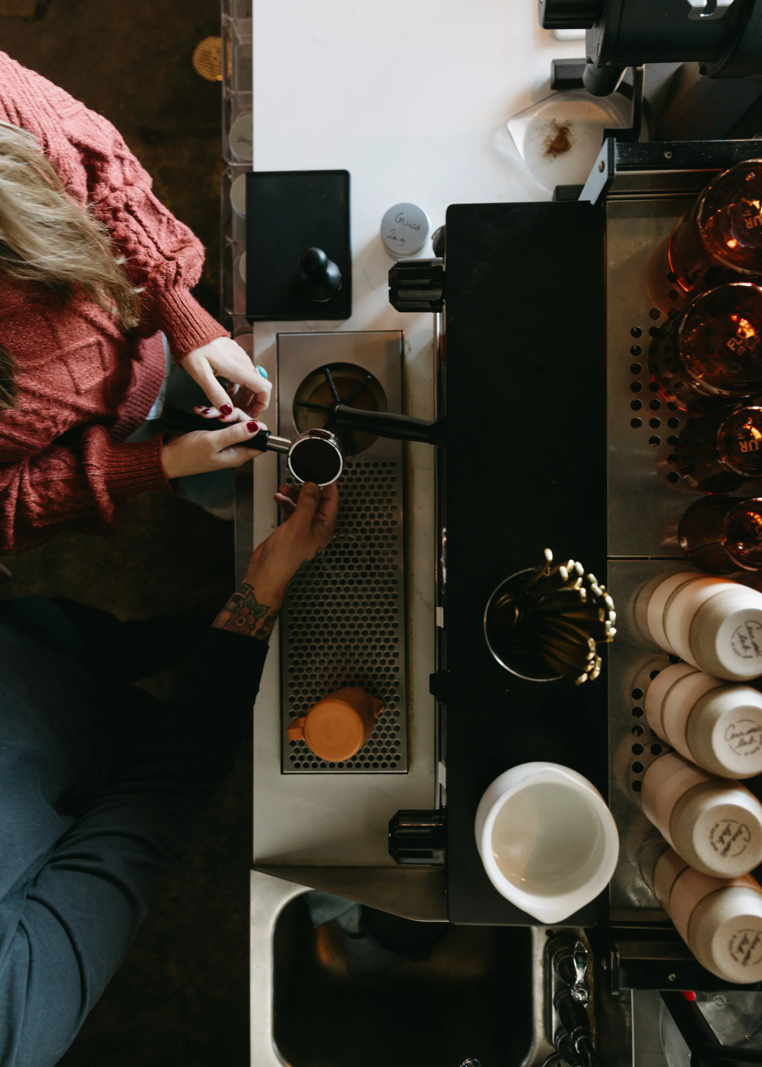 Two people making coffee using an espresso machine, with one person holding a portafilter and the other pressing a button, surrounded by coffee bottles and related equipment.