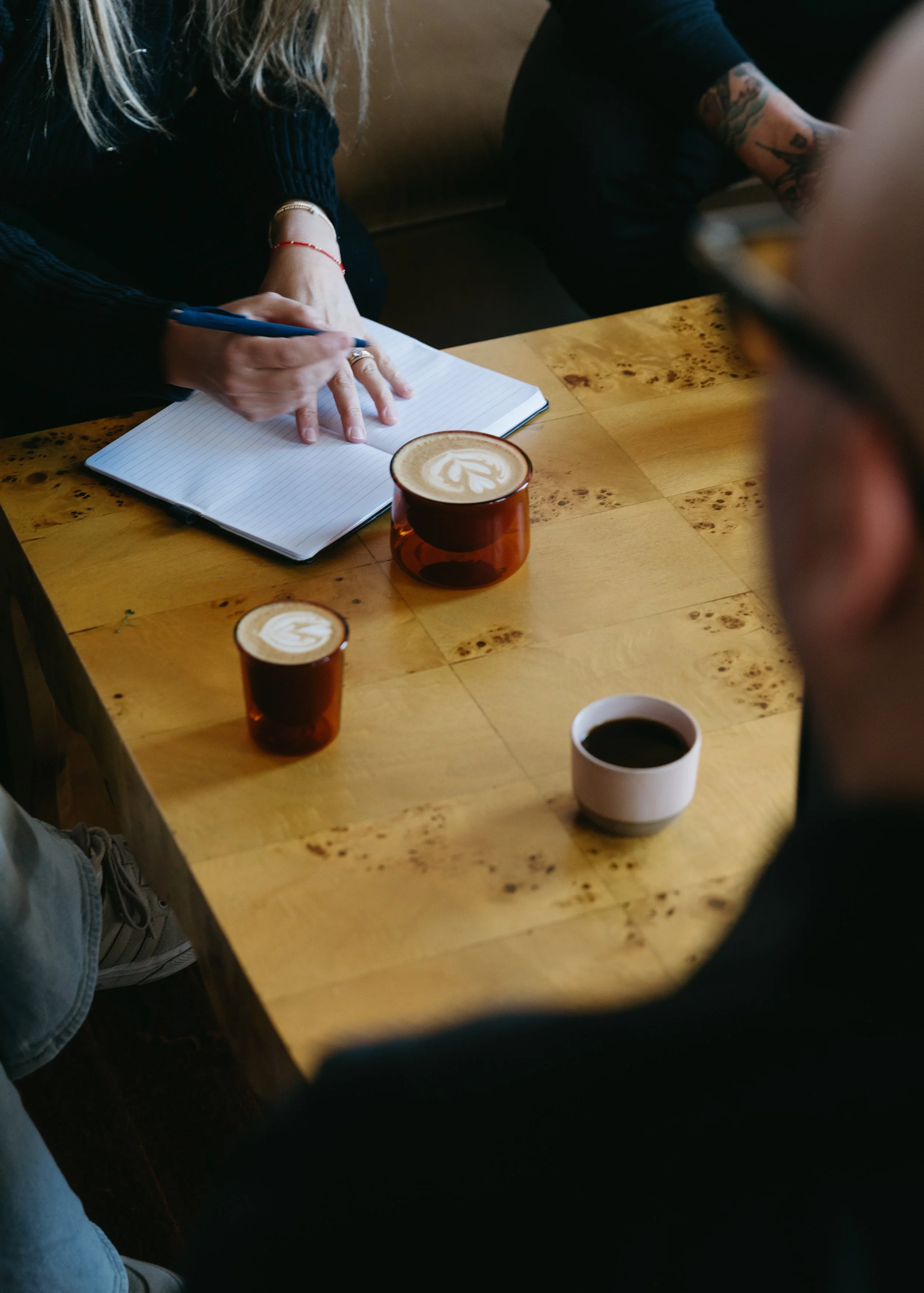 A person writing in a notebook at a wooden table, with two cups of latte art and a cup of black coffee in front of them, in a cozy cafe setting.