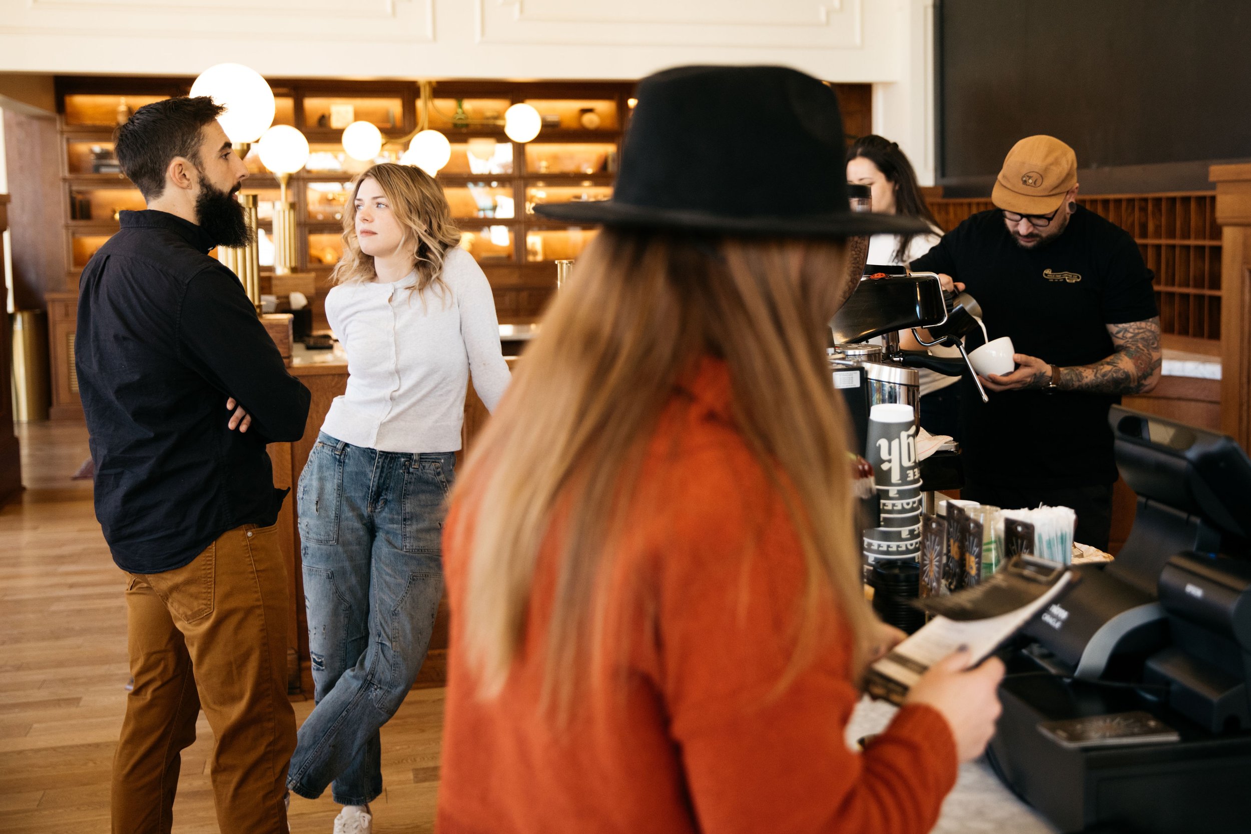 People in a coffee shop, with two customers talking, a barista preparing coffee, and another person reading a menu or receipt.