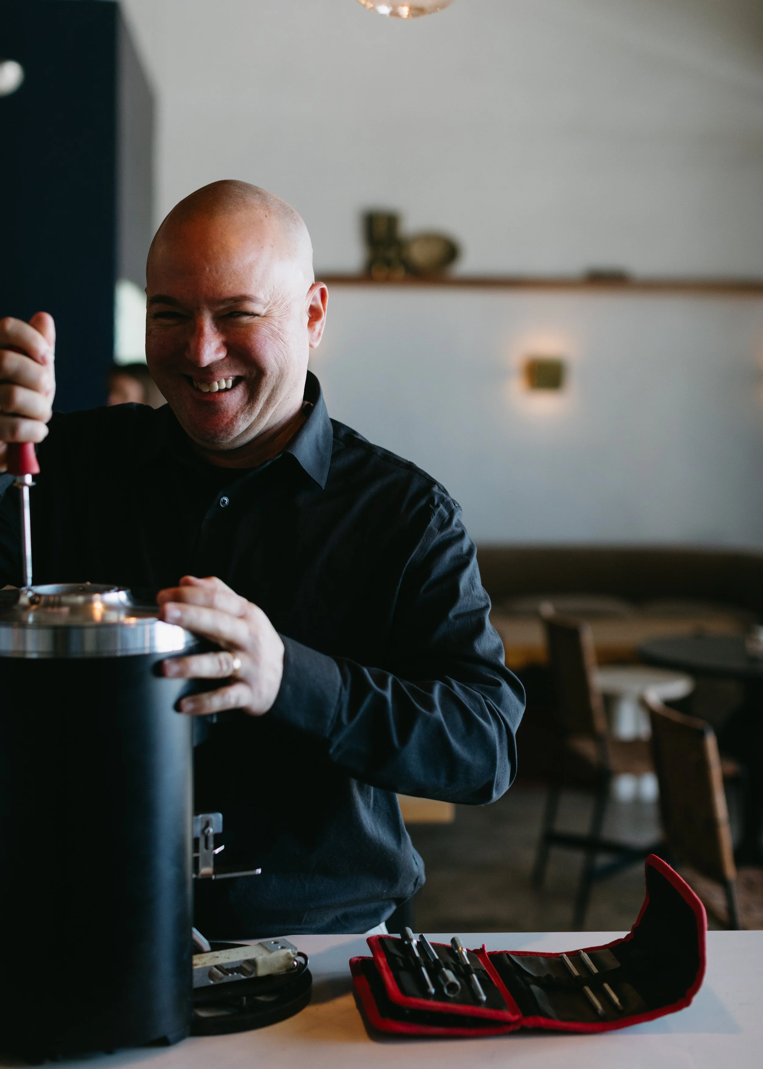 A smiling bald man working with tools in a well-lit room, possibly a workshop or café.