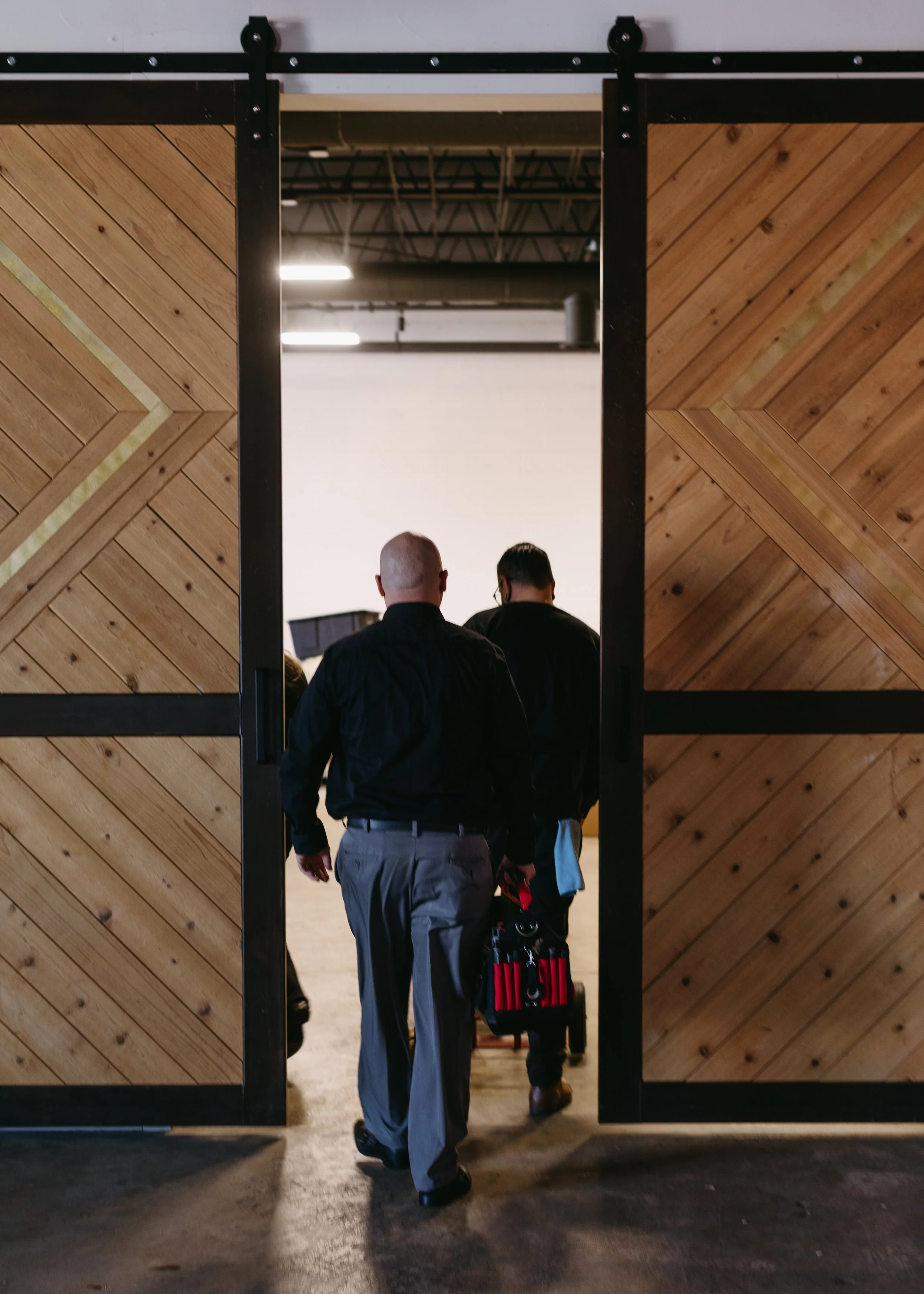 Two men entering through large wooden sliding barn doors into a brightly lit room, one carrying a red and black container.