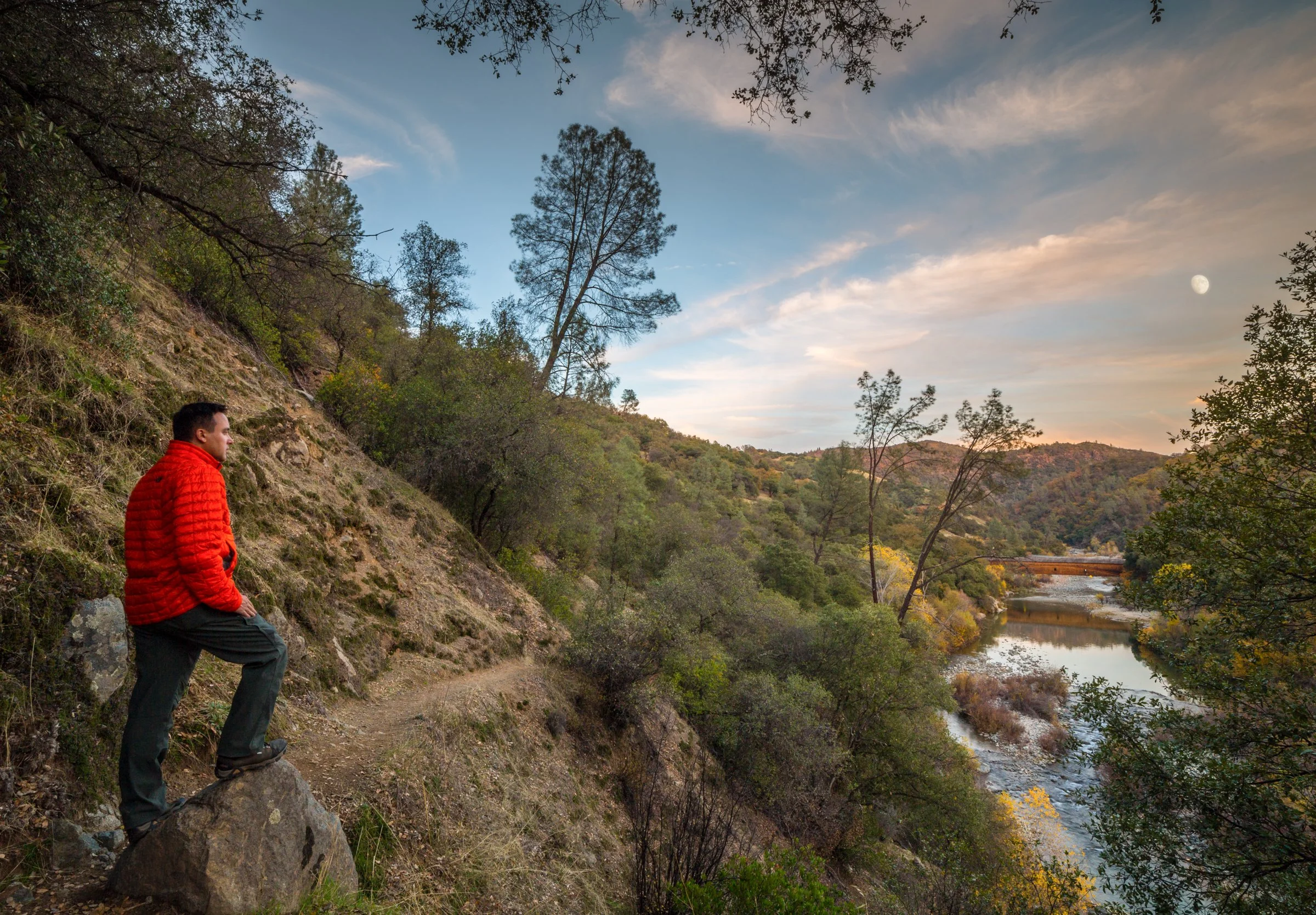 A man in a red jacket standing on a large rock by a trail, overlooking a river flowing through a hilly landscape dotted with trees, under a partly cloudy sky.
