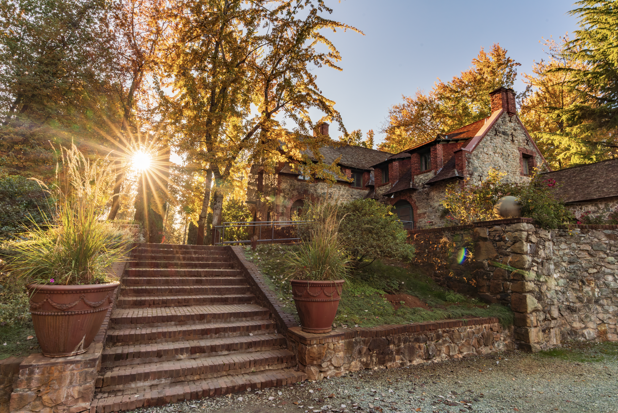 Sunset over a stone cottage with brick stairs, potted plants, and surrounded by colorful autumn trees.