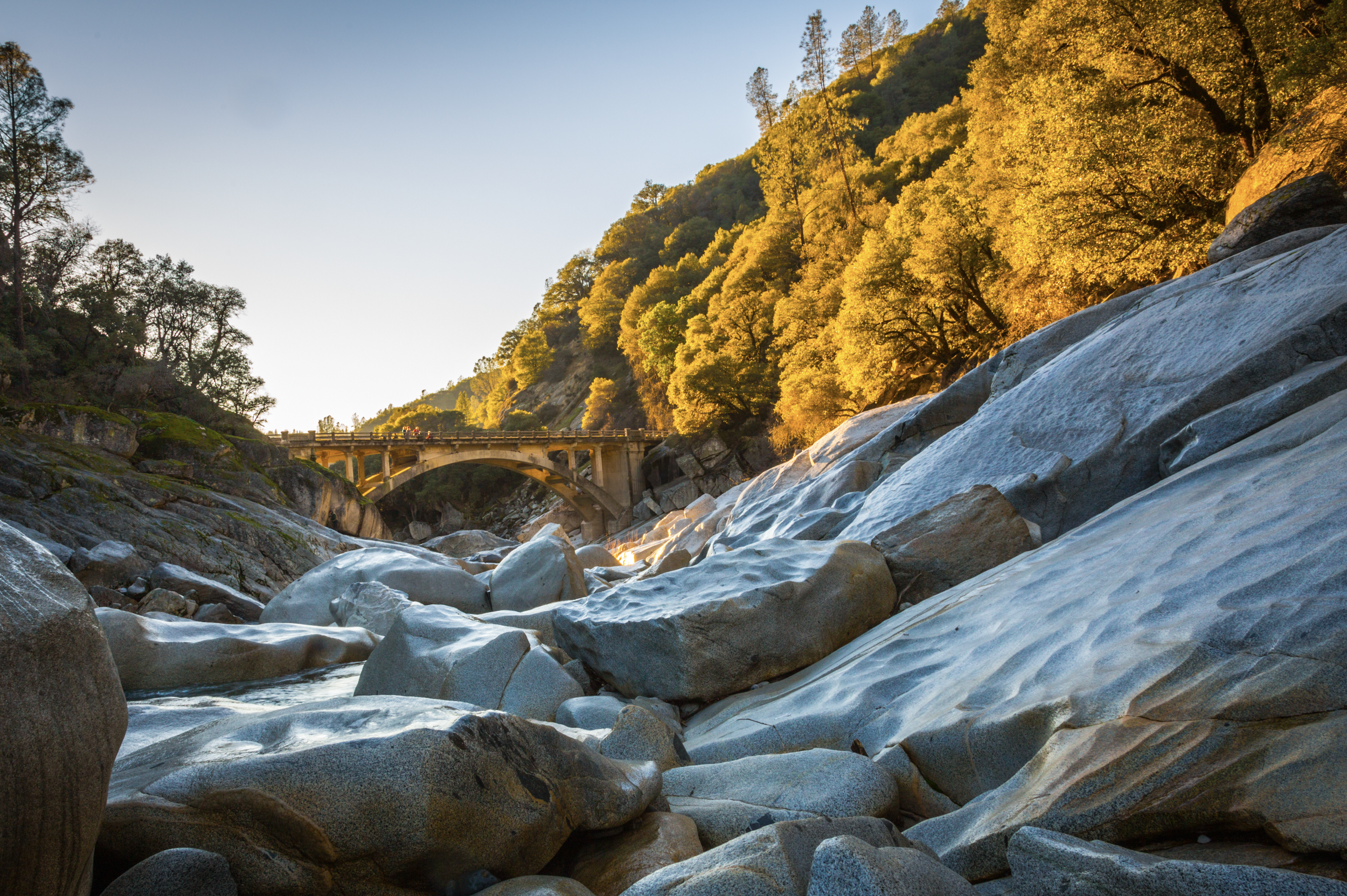 Sunlit riverbed with large rocks and a stone arch bridge in the distance, surrounded by trees with autumn foliage.