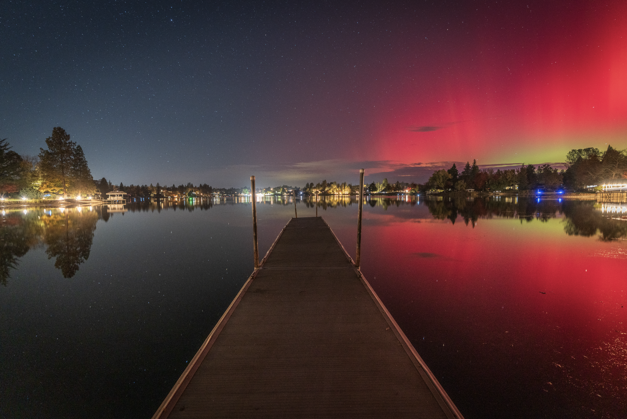A wooden dock extending into a calm lake at night, with colorful northern lights in the sky and trees along the shoreline reflected in the water.