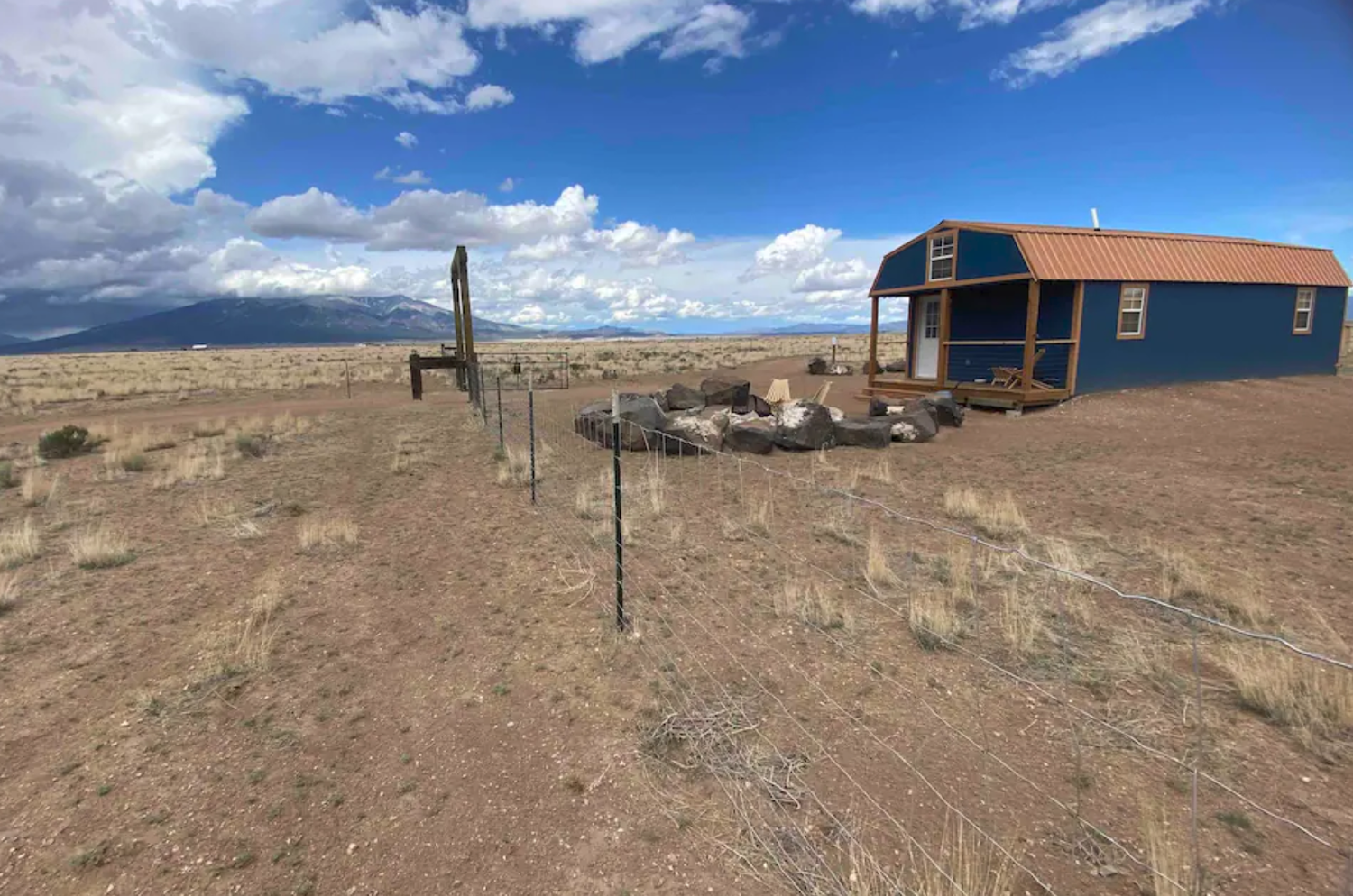 A small blue cabin with a brown roof on a dry, grassy plain under a partly cloudy sky. A wire fence runs alongside the cabin, and rocks are arranged in a circular pattern nearby. Distant mountains are visible in the background.