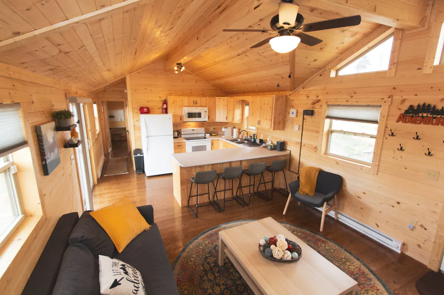 Cozy wooden cabin interior with a kitchen, living area, and high ceiling. Features include a sofa with cushions, a coffee table with decorative items, a kitchen island with stools, and windows with natural light. A ceiling fan and light fixture are visible, adding to the warm ambiance.
