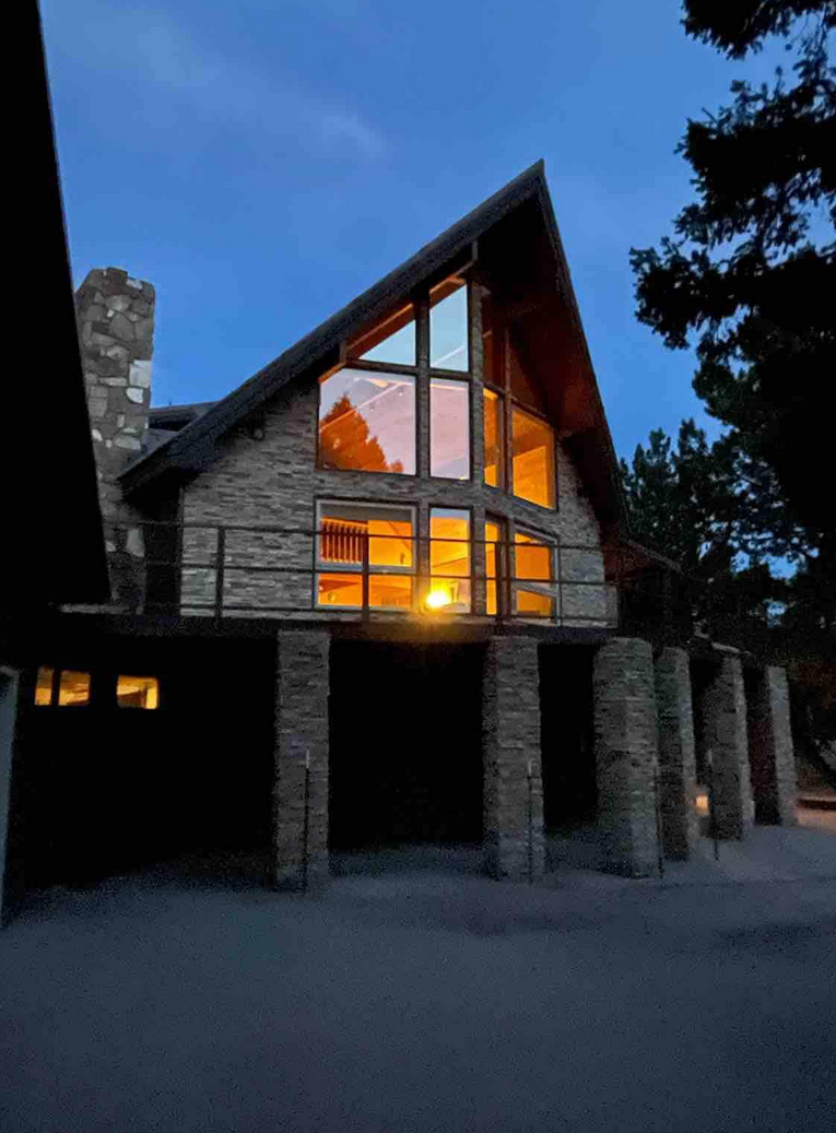 A-frame stone house with large windows illuminated at dusk