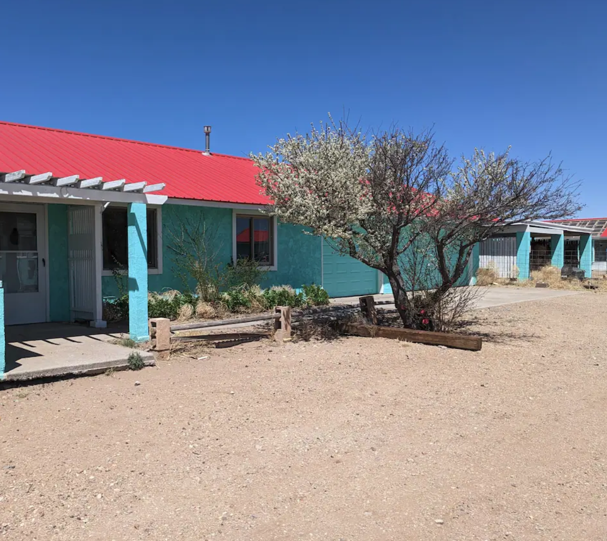 Single-story building with turquoise walls and a red roof in a desert landscape, featuring a tree and shrubbery in the foreground.