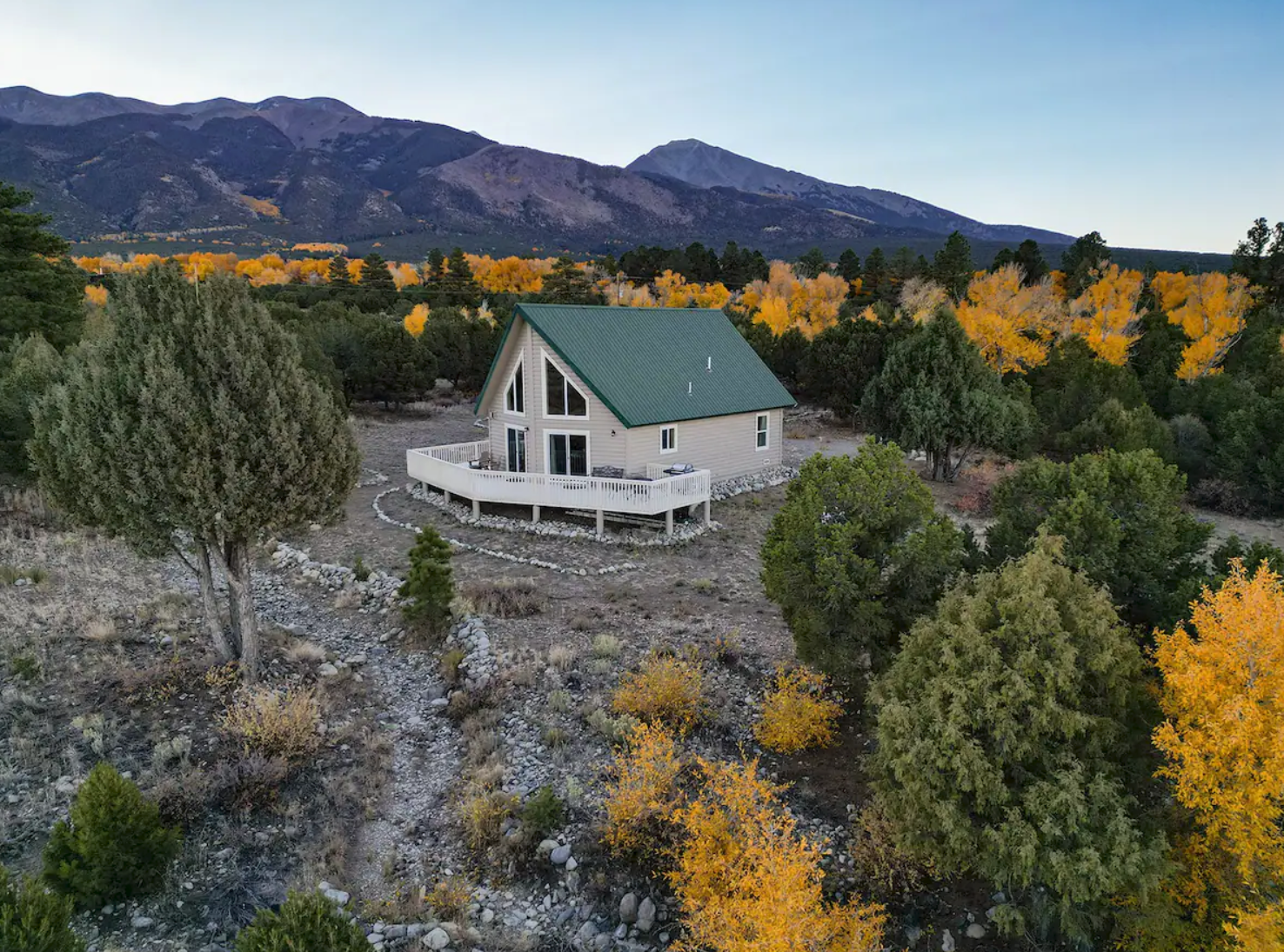 A-frame cabin with a green roof surrounded by autumn trees and mountains.