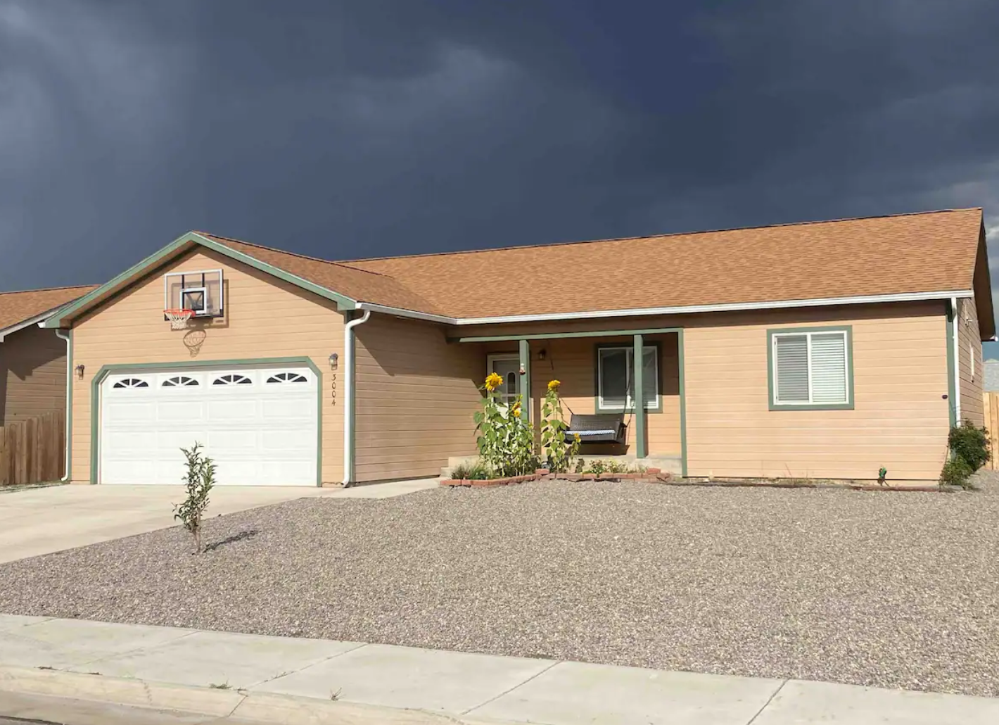 Single-story tan house with a basketball hoop on the garage, gravel front yard, and sunflowers by the porch.