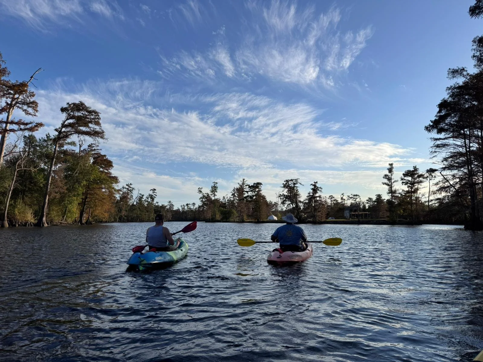 Two kayakers paddling across a scenic Southwest Louisiana waterway during a Bayou Glamping Co. outdoor adventure, surrounded by cypress trees and blue skies.