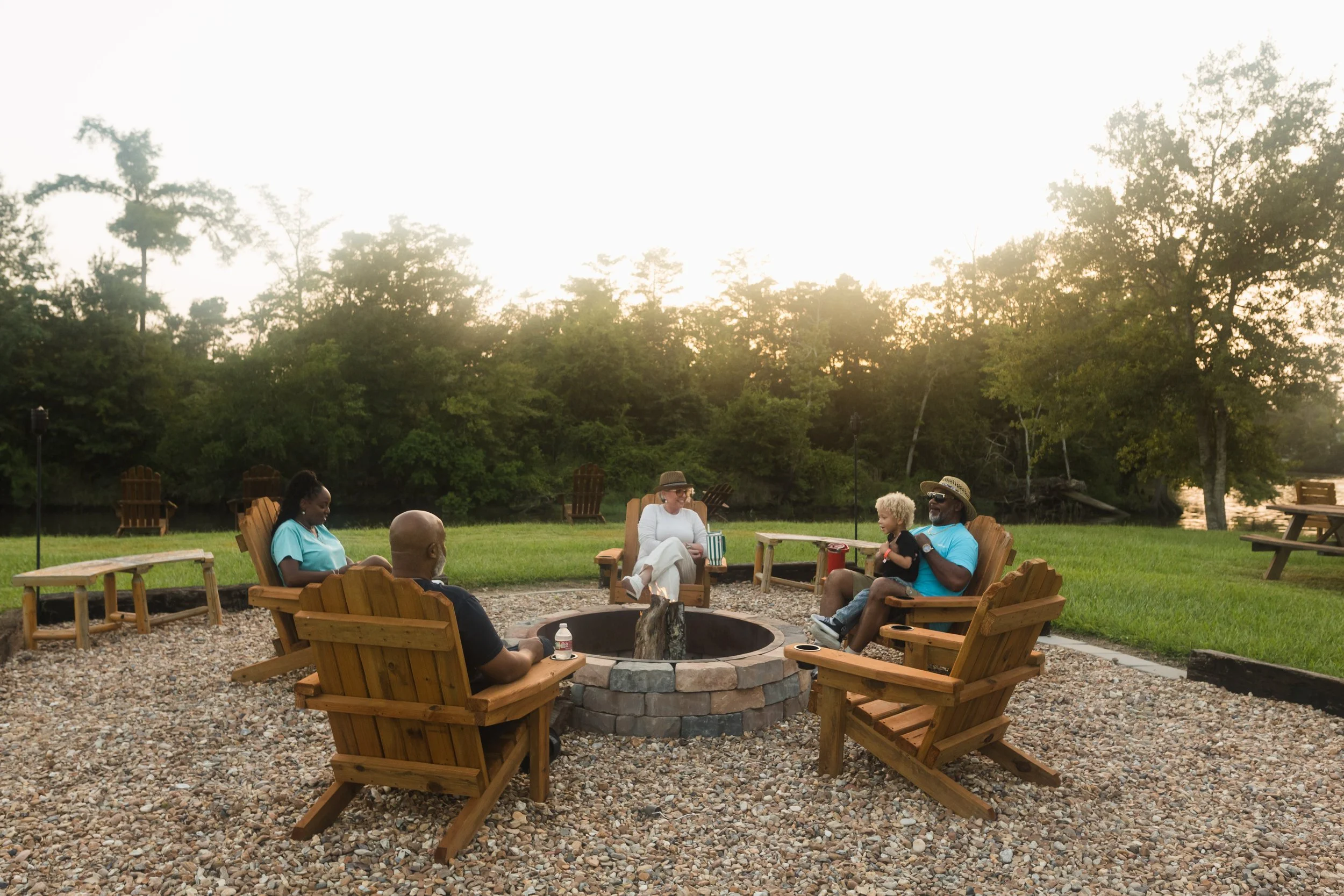 Group of people sitting around Bayou Glamping Co's upscale fire pit outdoors on a gravel surface, with trees and the Calcasieu River in the background during sunset.