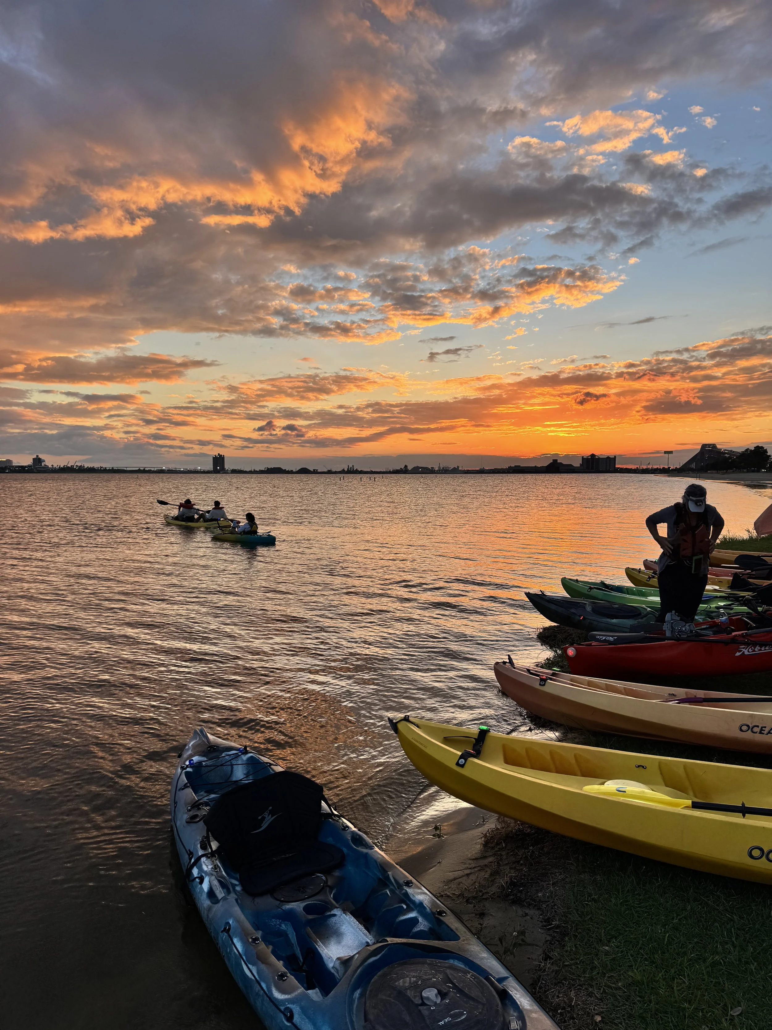 A smiling young man and woman sitting on a rug outside a beige canvas tent, enjoying camping in the sunlight, with a wooden deck and camping trailer in the background at Bayou Glamping Co. in Southwest Louisiana
