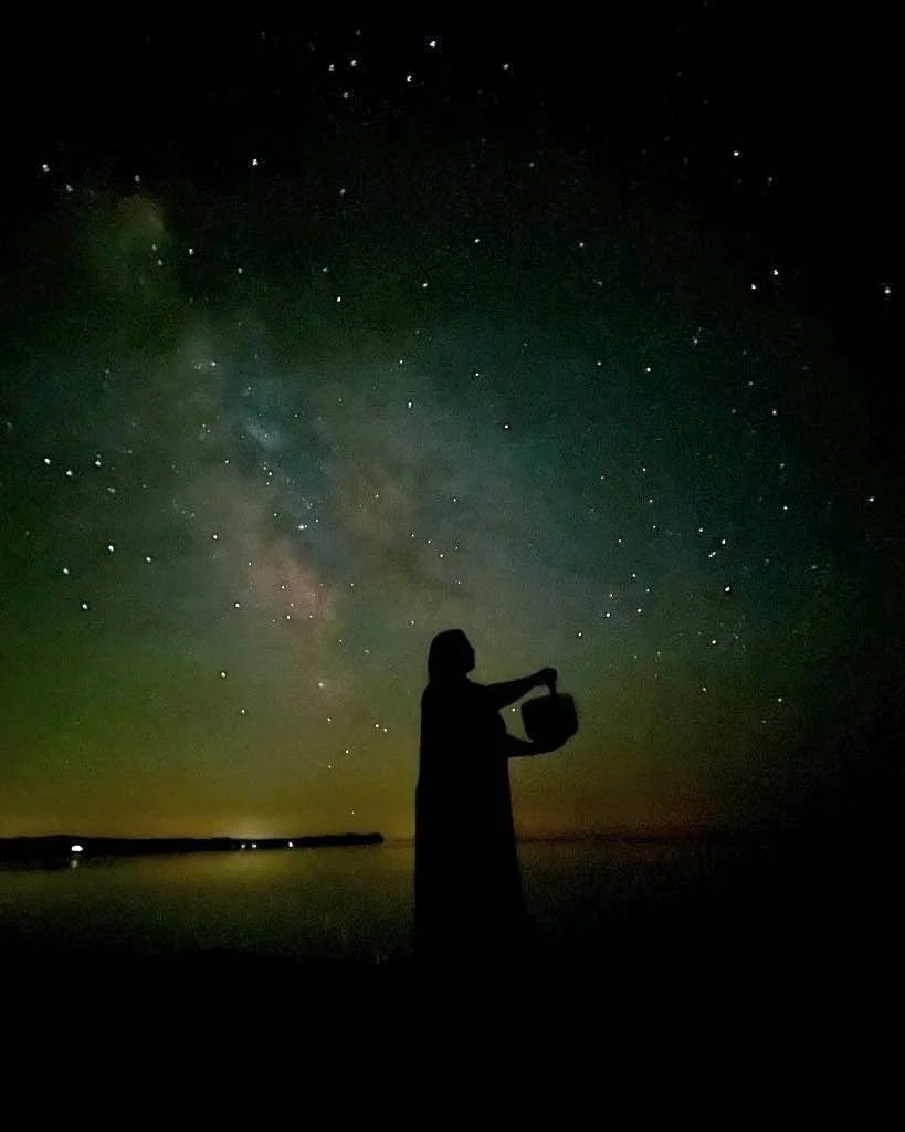 Silhouette of a person standing by water holding a bucket under a starry night sky with the Milky Way galaxy visible.
