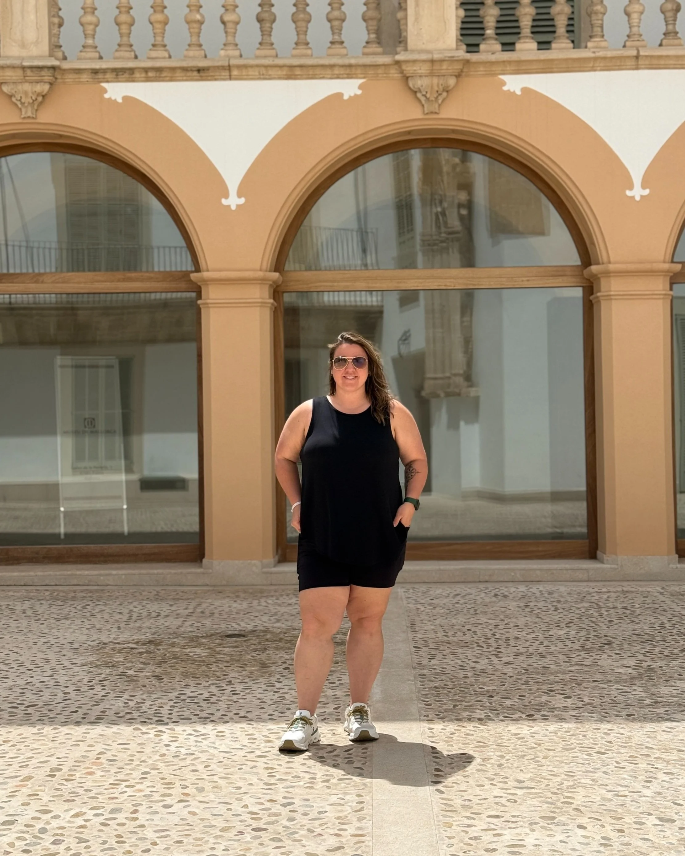 A woman standing outside in front of a building with large arched windows, wearing sunglasses, a black sleeveless top, and black shorts, smiling with her hands in her pockets.