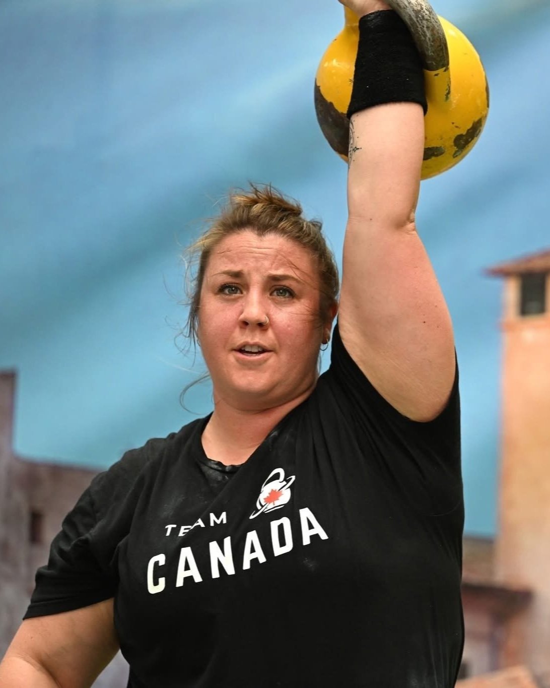 A woman wearing a black shirt with a Canada logo lifting a yellow and black kettlebell overhead during a workout outdoors with cloudy sky and buildings in the background.
