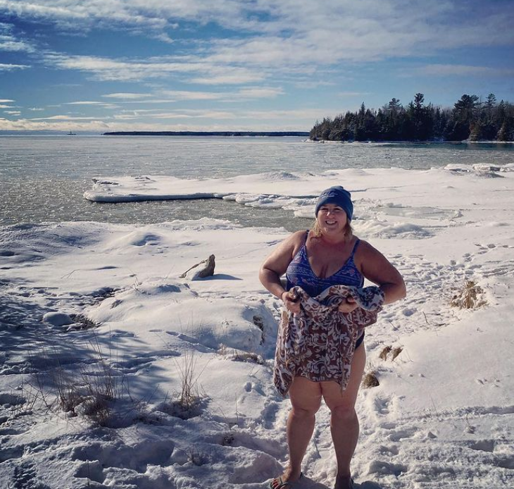 Woman in a blue swimsuit and beanie standing on snow-covered shore holding a towel, with icy water and trees in the background.
