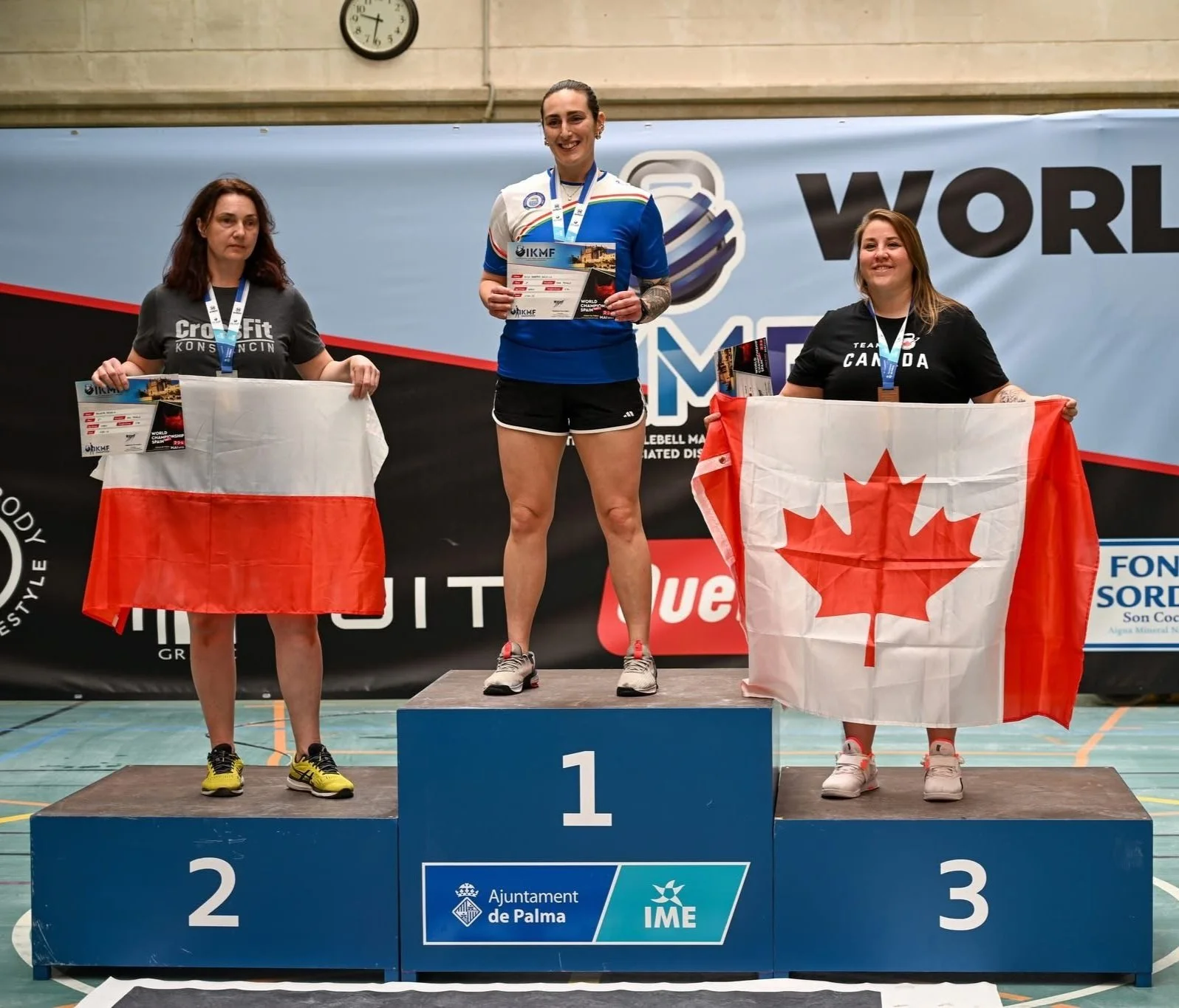 Three women standing on a winners' podium after a sports competition. The woman in the center on the first place holds a certificate and medals, wearing a blue shirt and black shorts. The woman on the left on the second place holds a Polish flag and wears a dark t-shirt and shorts. The woman on the right in third place holds a Canadian flag and wears a black t-shirt and shorts. They are indoors with a blue background and a clock on the wall.