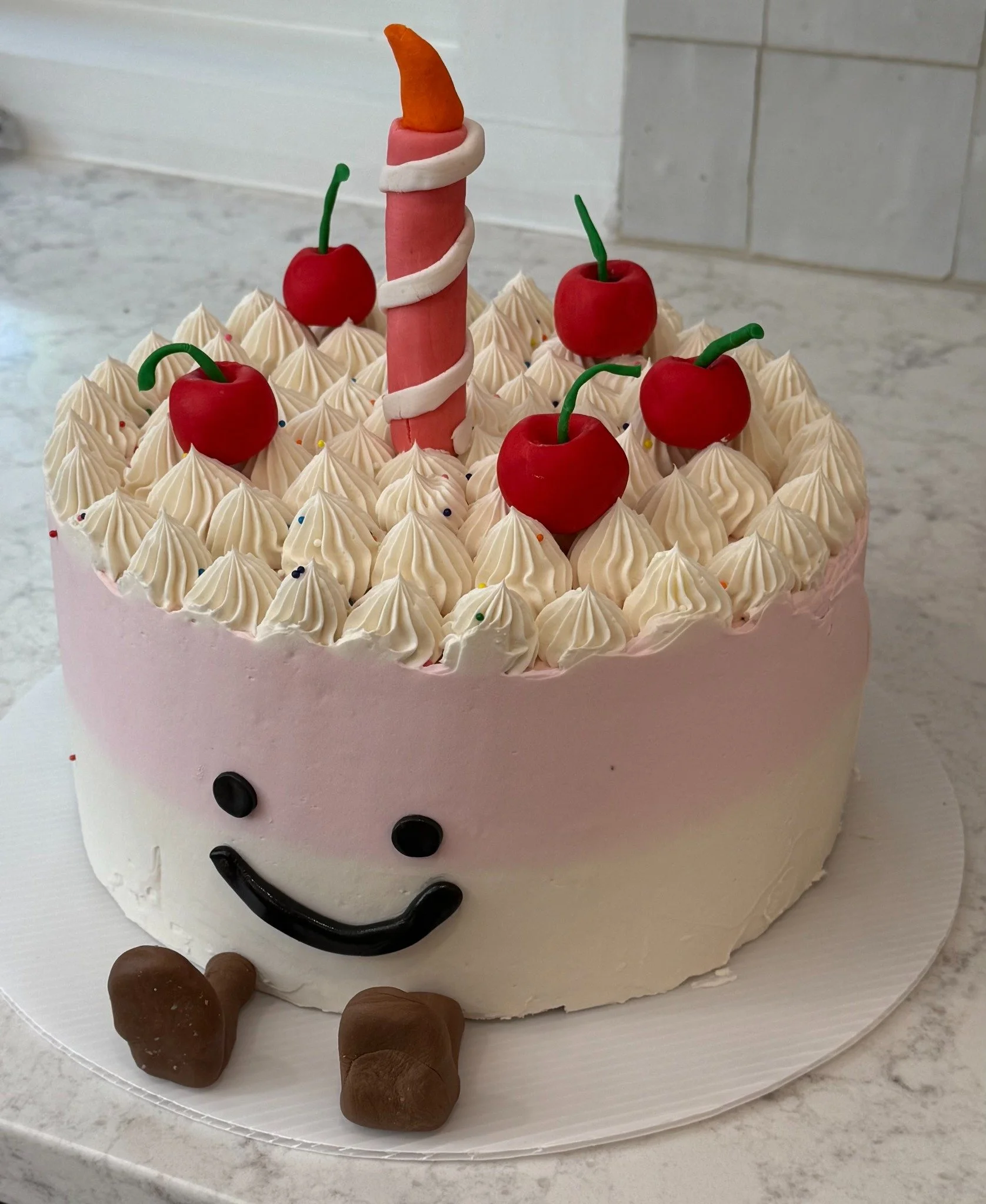 Smiling pink-and-white birthday cake with cherry decorations and a handmade candle topper. A past custom cake for inspiration.
