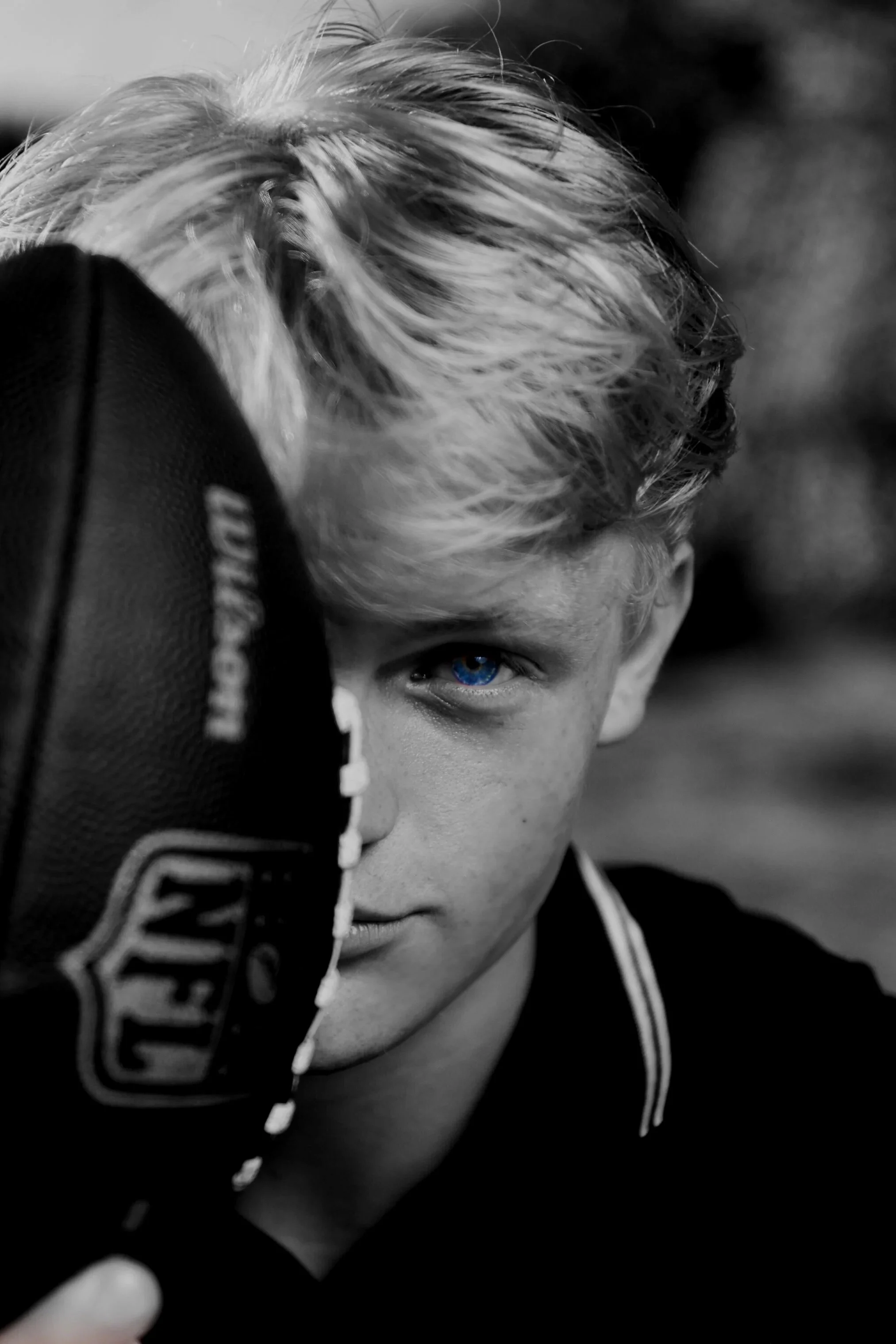 A black and white portrait of a young person with blonde hair and striking blue eyes, partially obscured by a black baseball glove held up close to their face.