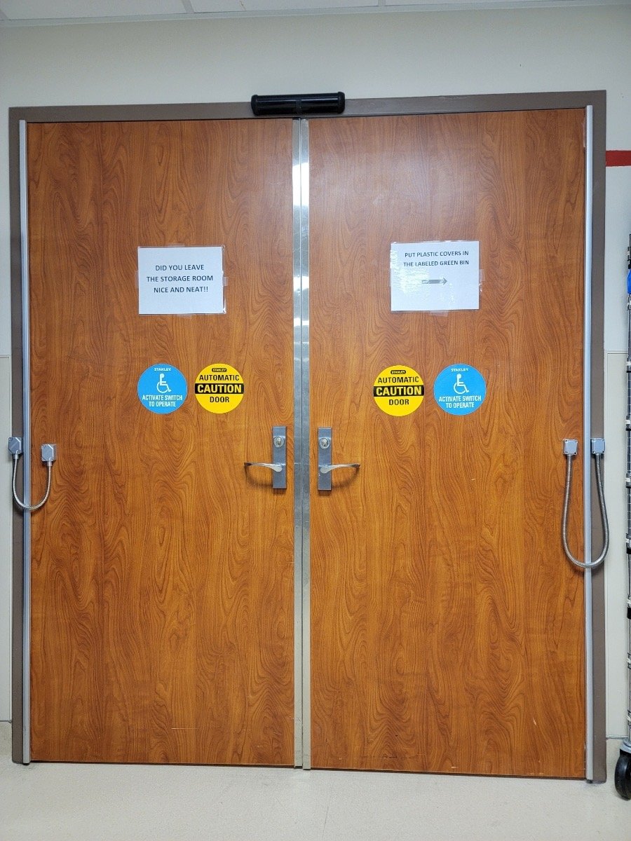 Wooden double doors with silver handles, featuring signs "Do you leave the storage room nice and neat?!!" and "Put plastic covers in the labeled green bin." Also, blue wheelchair symbols and yellow "Automatic Caution Door" stickers.