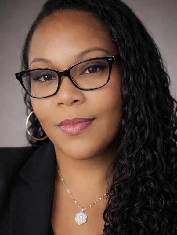 Professional woman with glasses, hoop earrings, and a necklace, wearing a black blazer, with curly hair, looking at the camera.