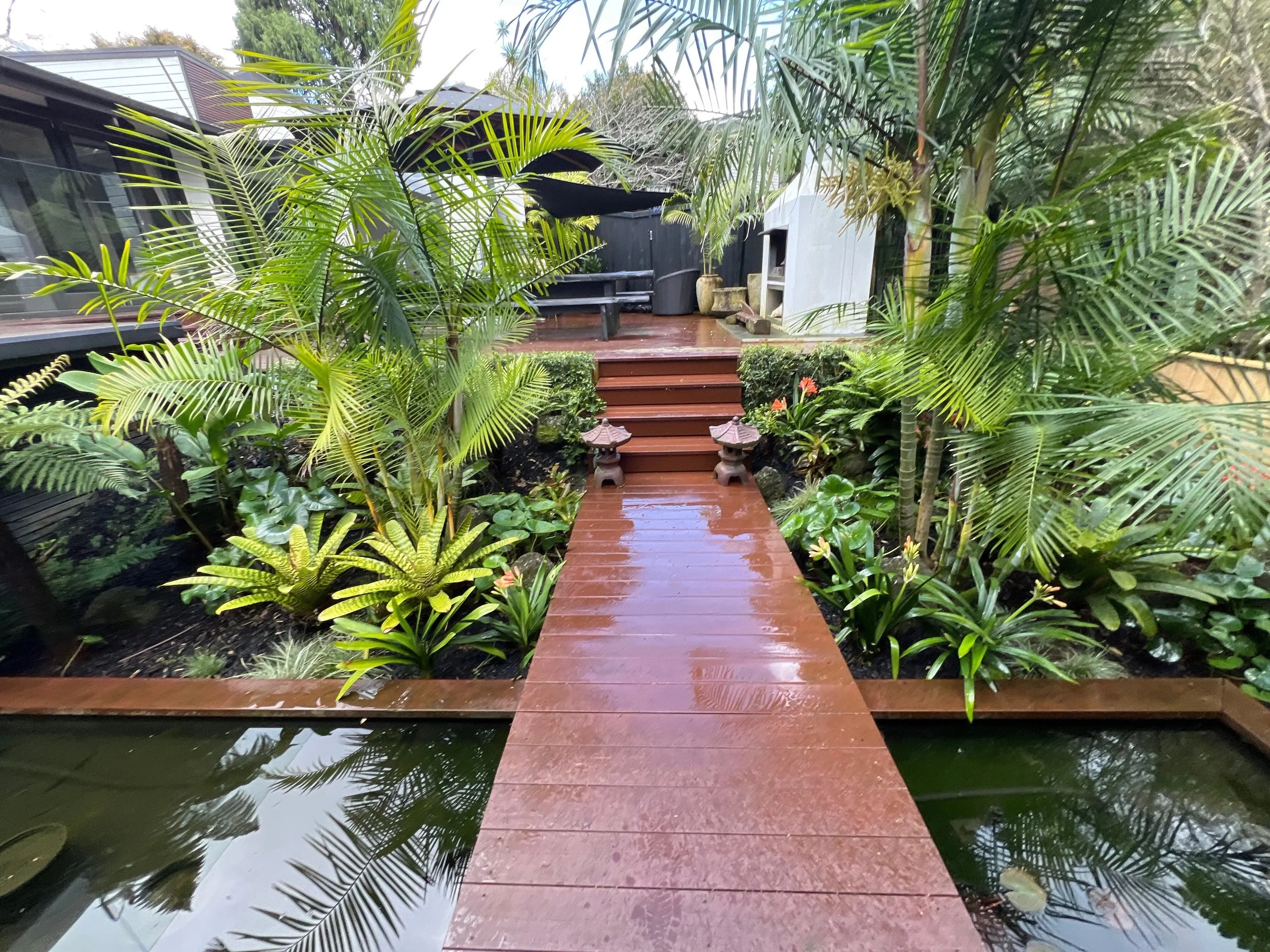 Rain-soaked wooden pathway through a lush tropical garden, leading to a deck with outdoor furniture and a built-in grill.