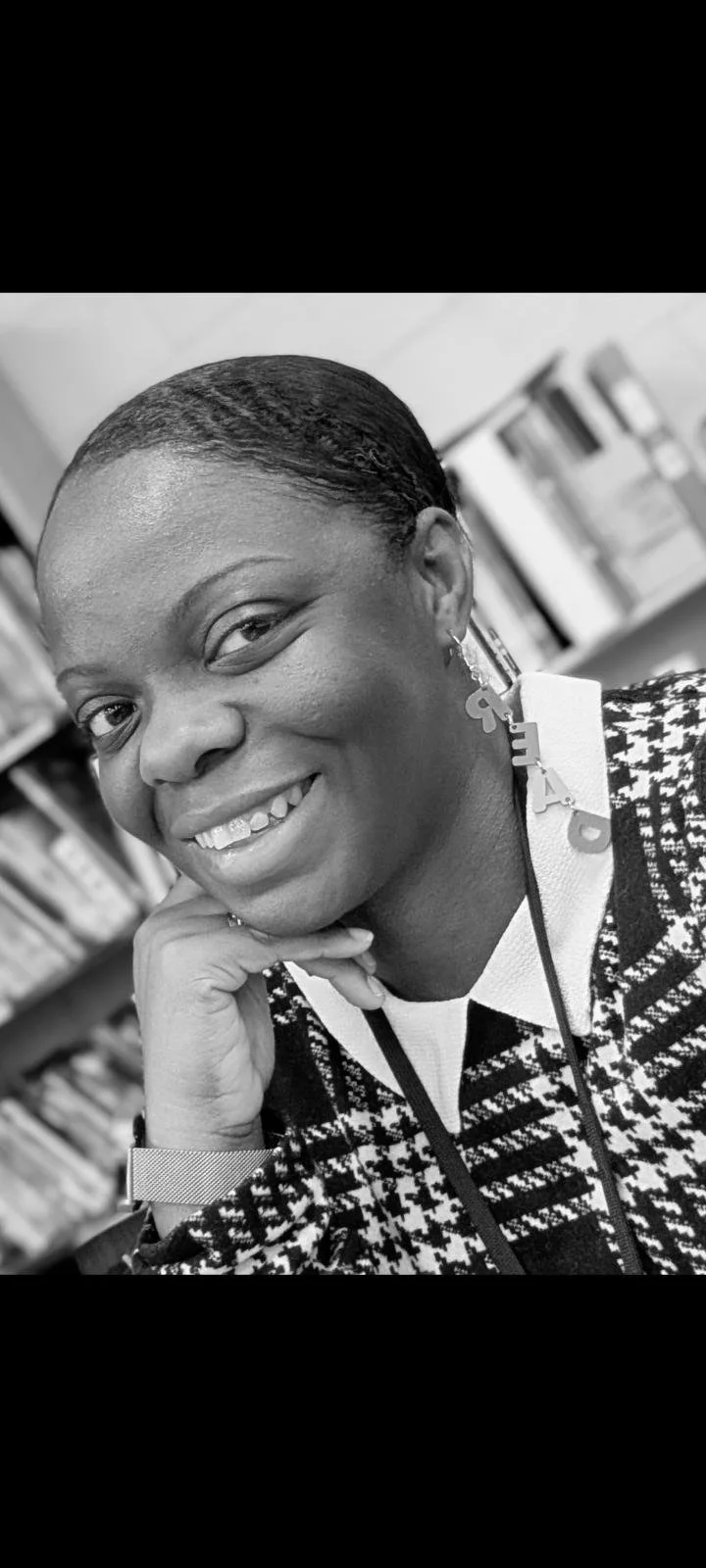 Black and white close-up of a smiling woman with smooth, short hair, wearing earrings, a patterned sweater, and a lanyard with an ID badge, in a library or bookstore setting.
