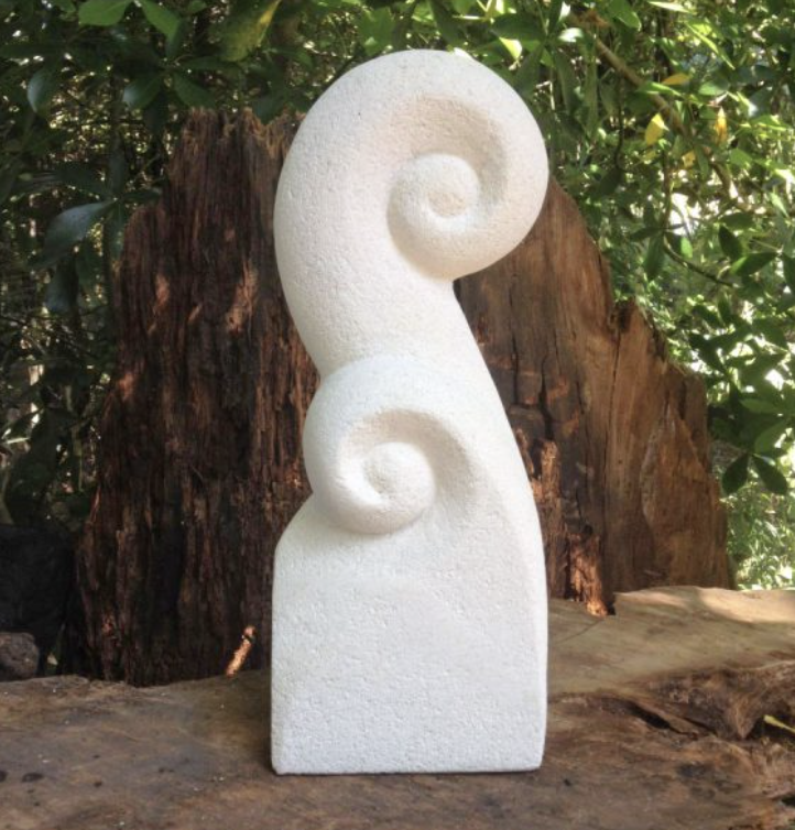 A white sculptural art piece with twin spiral shapes, placed outdoors on a wooden surface against a background of green foliage and a tree trunk.
