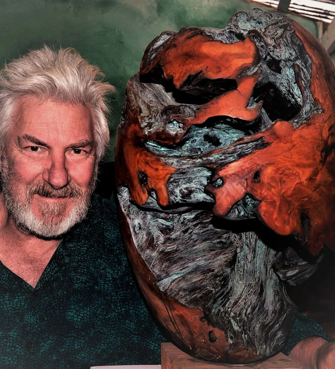 A man with gray hair and a gray beard smiling, standing next to a large, intricately carved wooden sculpture of a face.