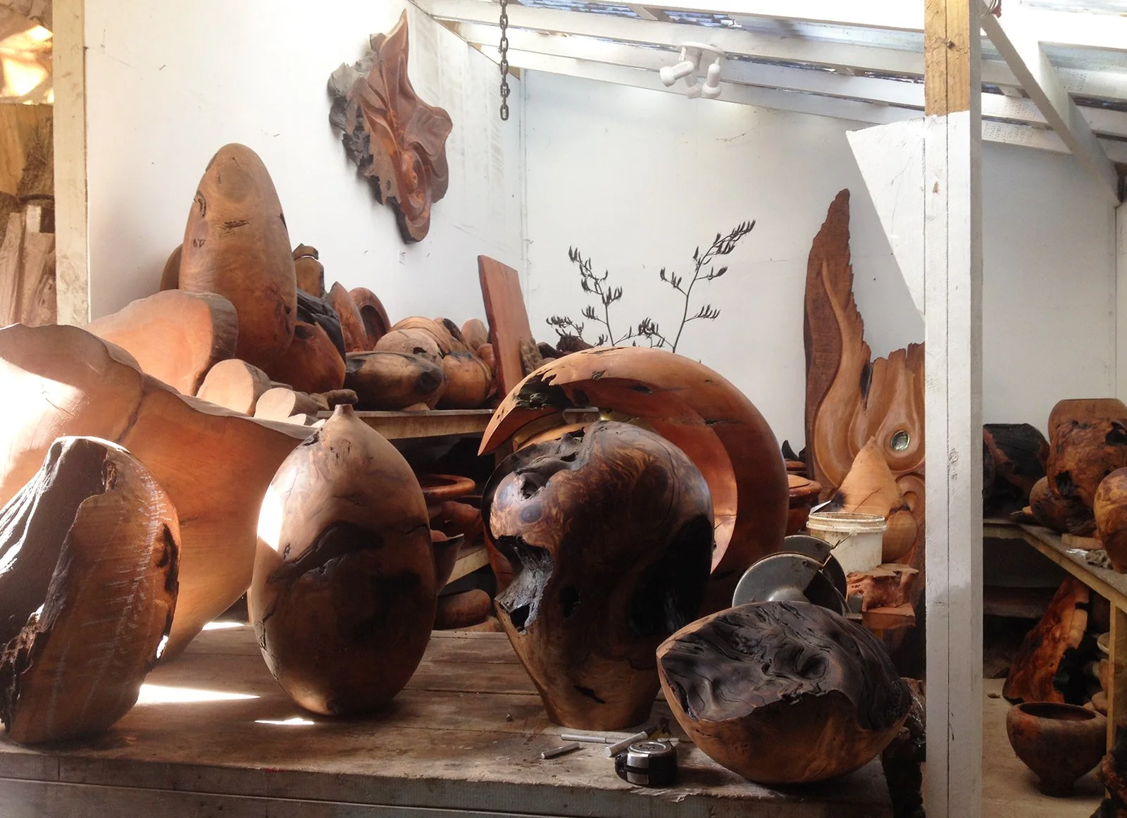 Various wooden bowls, vases, and sculptures displayed on shelves and tables in a workshop, with some sunlight coming through openings in the ceiling
