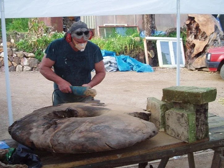 A person wearing a mask, sunglasses, and a dark T-shirt is carving or shaping a large piece of wood outdoors under a canopy. The workspace includes large blocks of stone and wood, with a background featuring various tools, equipment, and natural elements.