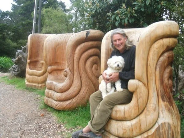 An artist sitting on a large wooden sculpture holding a small white dog. The sculpture is carved from a Kauri tree trunk with swirling patterns. There are similar sculptures lined up behind him, and trees and greenery in the background.