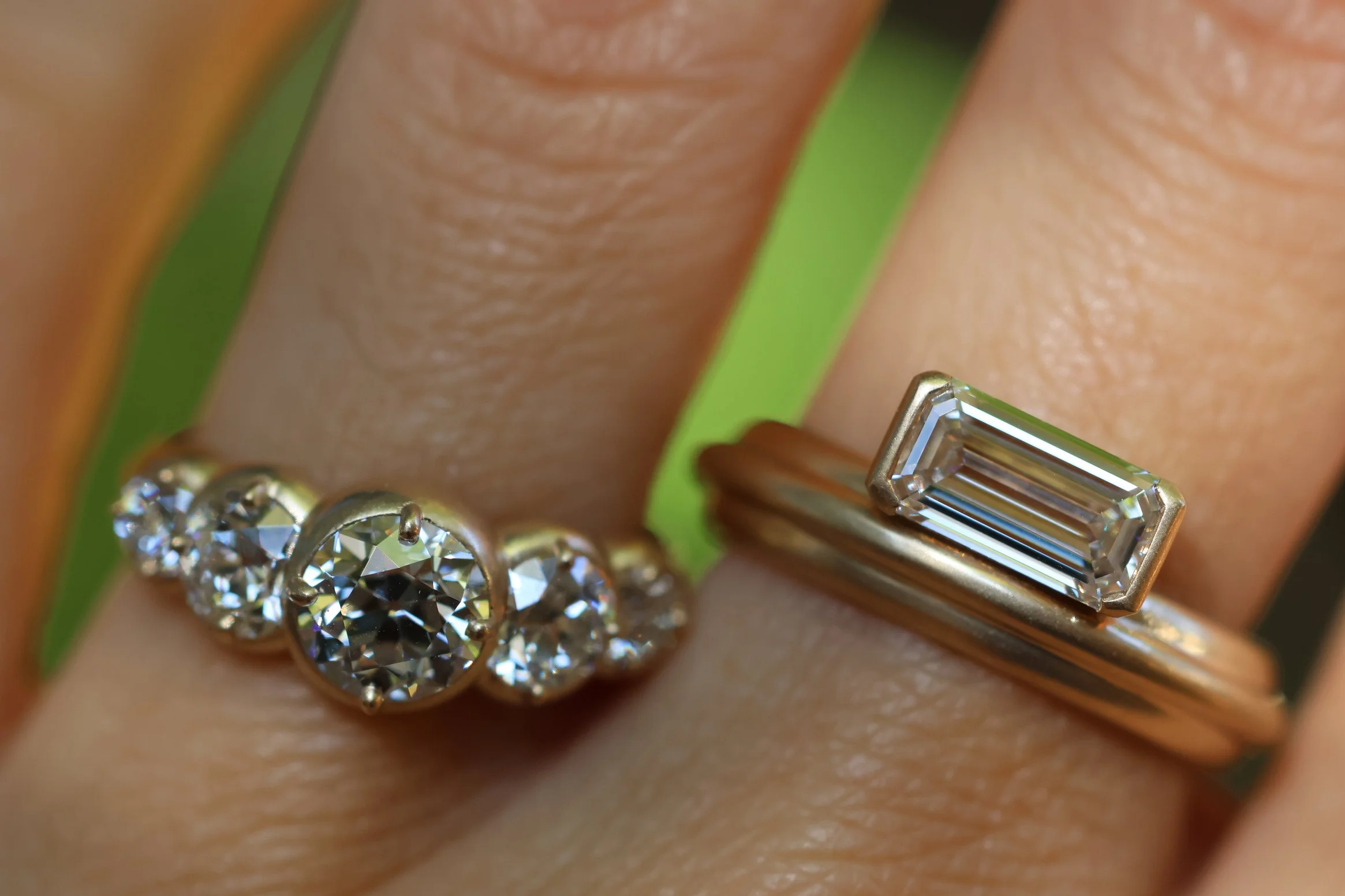 Close-up of two rings on fingers: one with graduated old mine cut diamonds and a central larger diamond, and the other with a rectangular baguette-cut diamond.