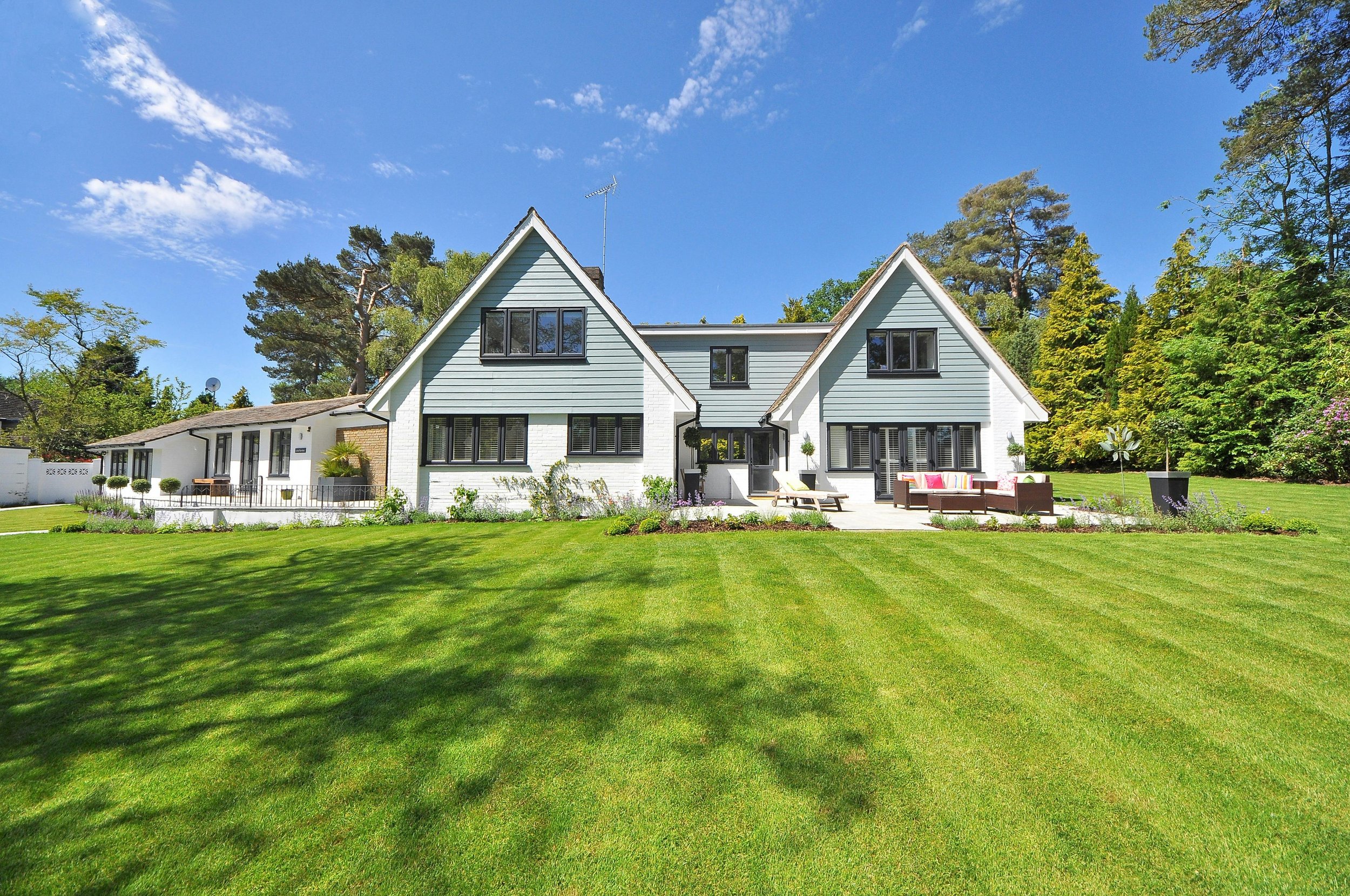 Large white and blue house with a spacious green lawn and surrounding trees under a clear blue sky.