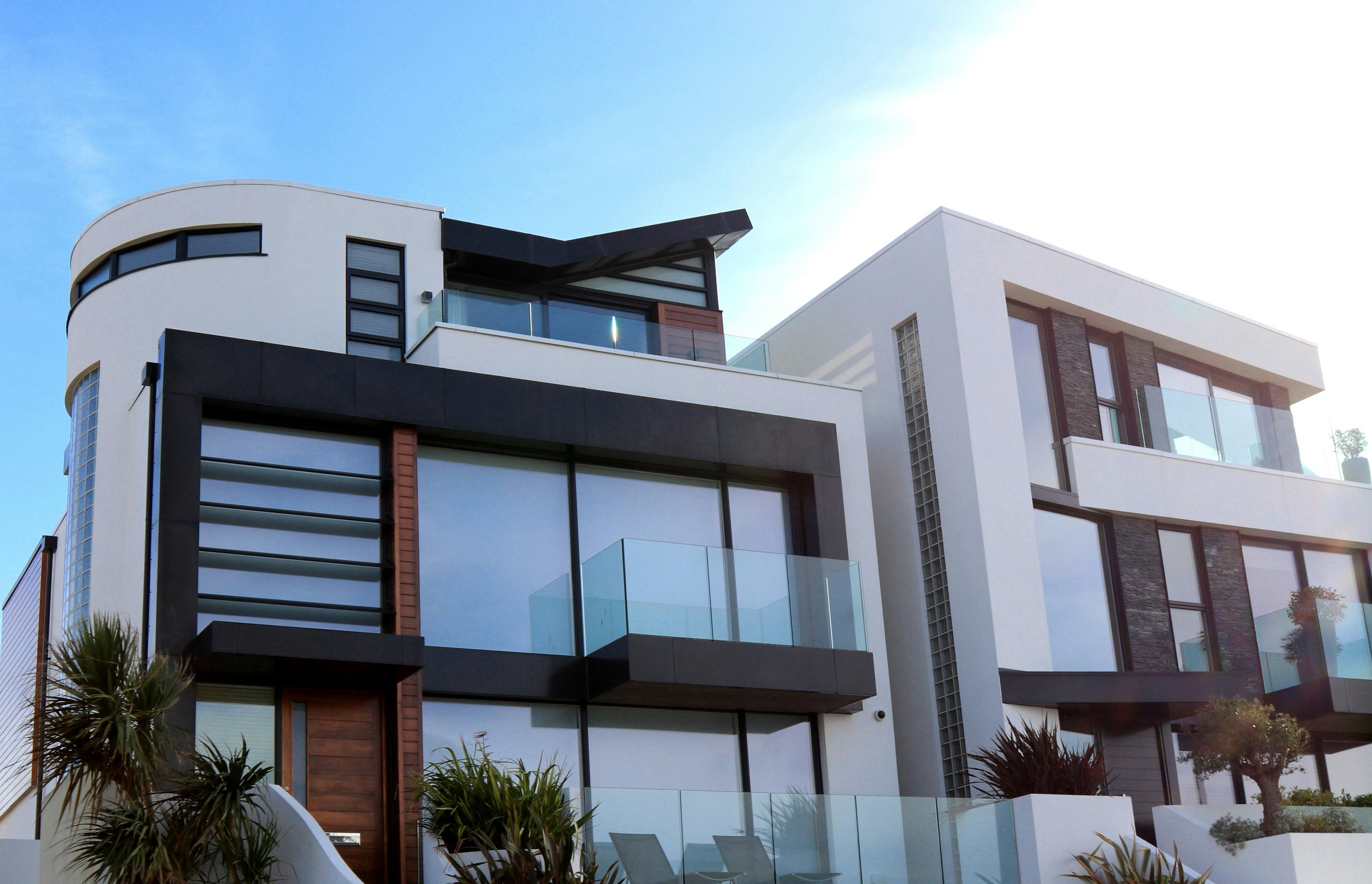 Modern white and black two-story house with large glass windows, balconies, and minimalist landscaping under a clear blue sky.