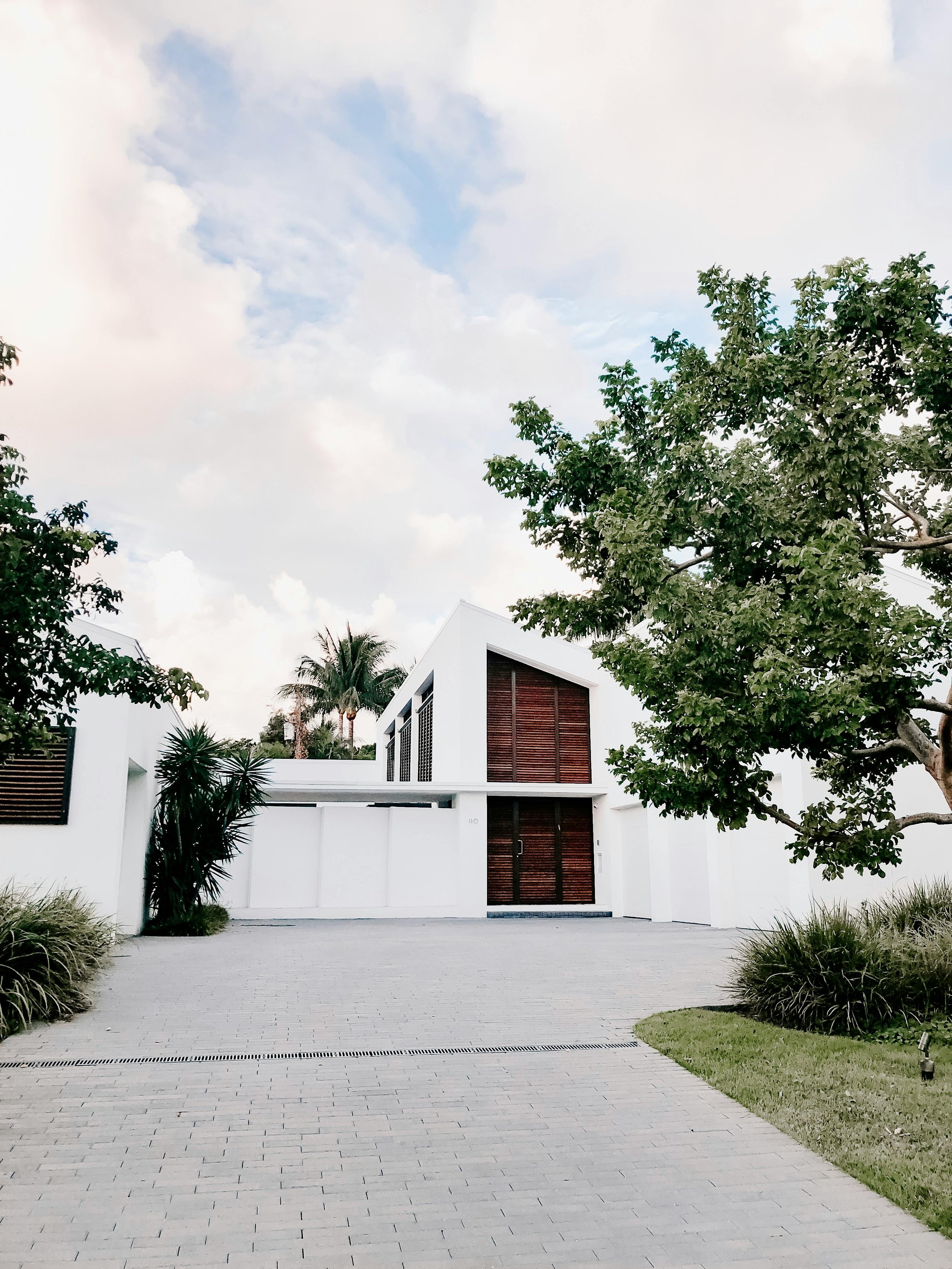 Modern white house with large wooden doors, surrounded by greenery and a paved driveway under a partly cloudy sky.