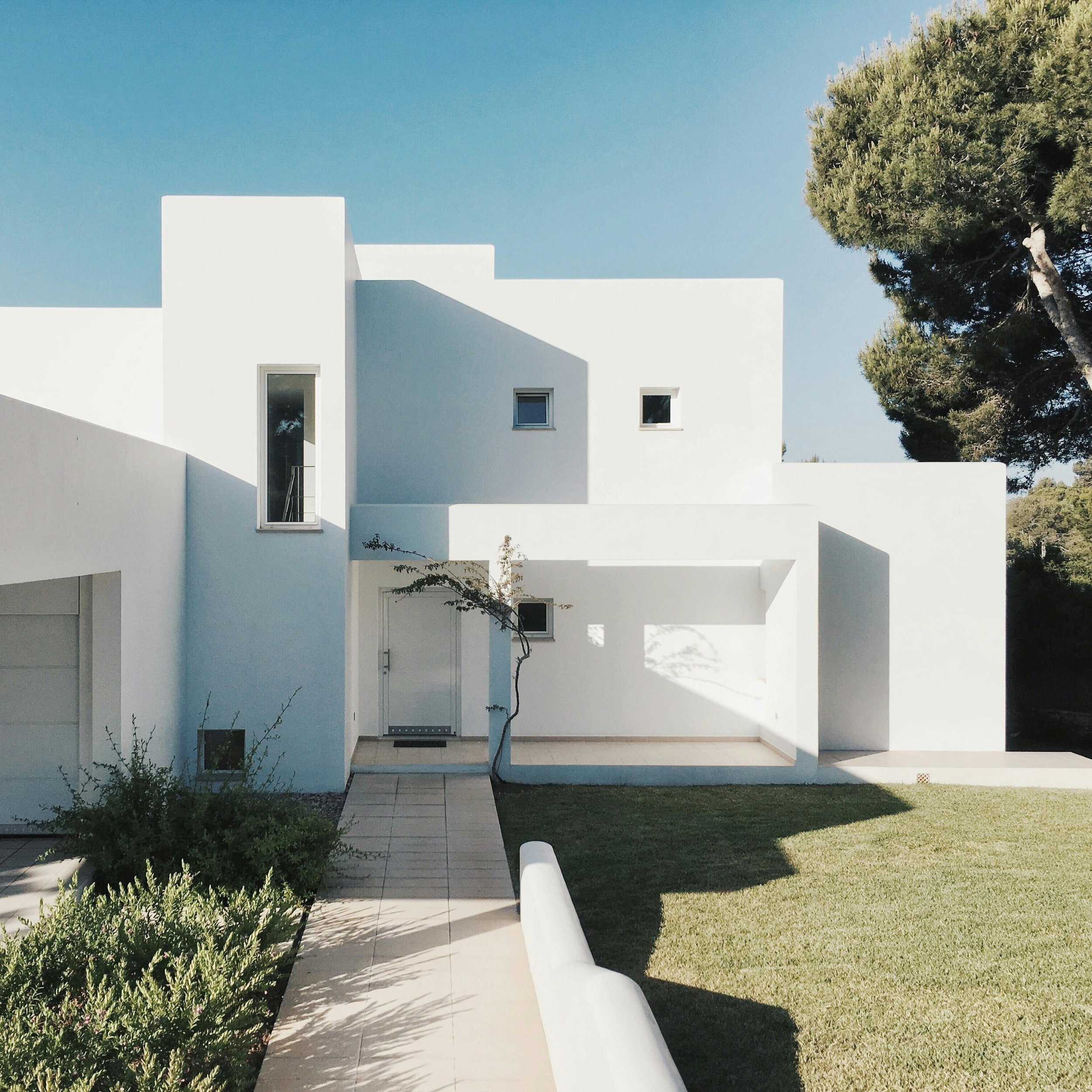 Modern white minimalist house with geometric architecture, surrounded by greenery and a tree, under a clear blue sky.