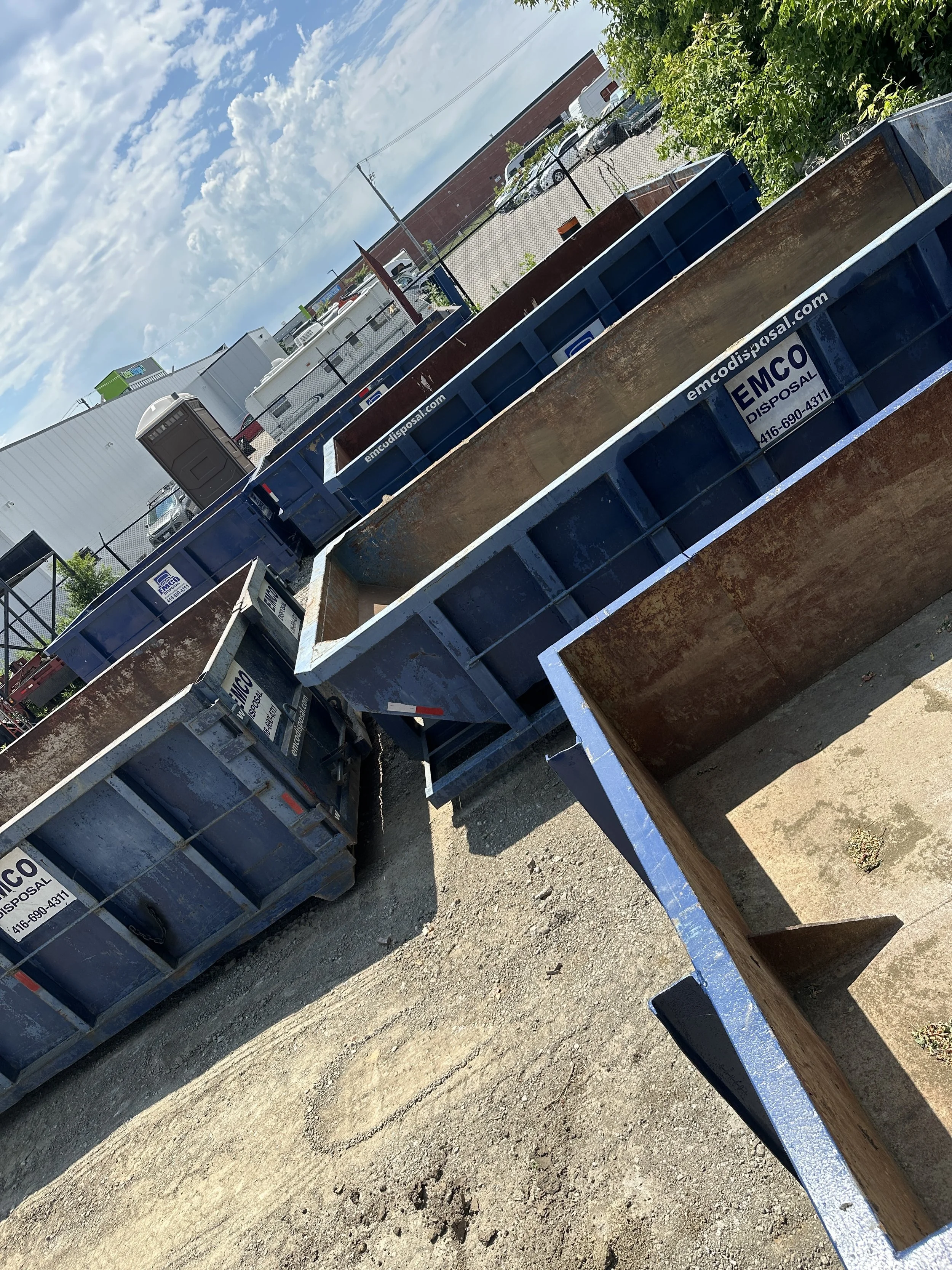 Industrial blue dumpsters on a gravel lot with buildings and trees in the background.