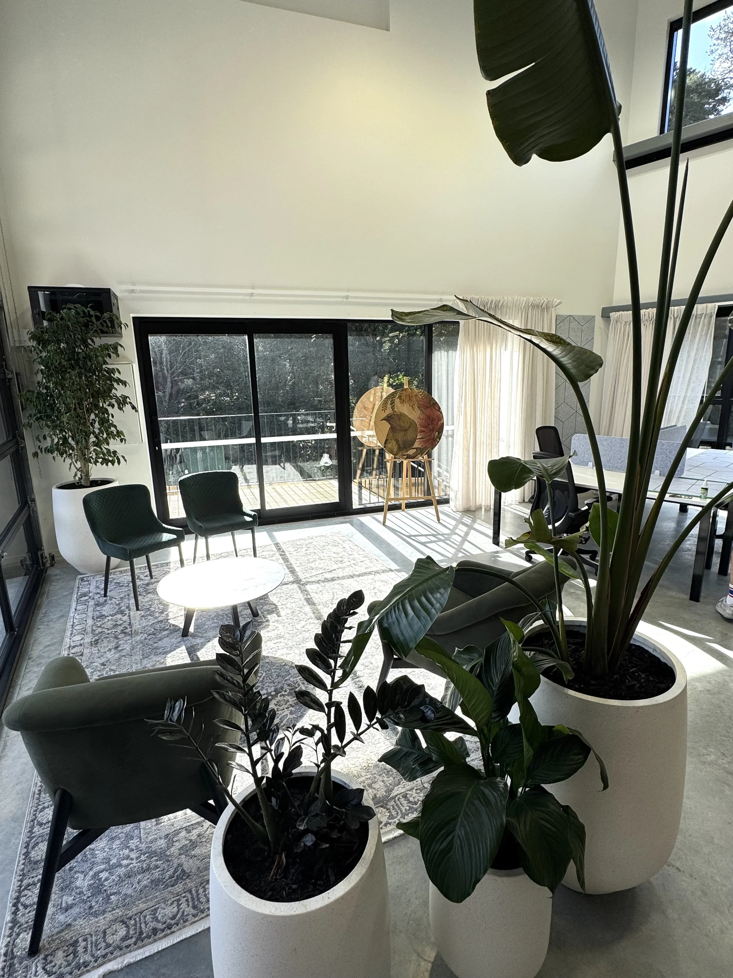 Modern open-plan interior living space with large windows, green armchairs, a round black coffee table, plants, and a kitchen area in the background.