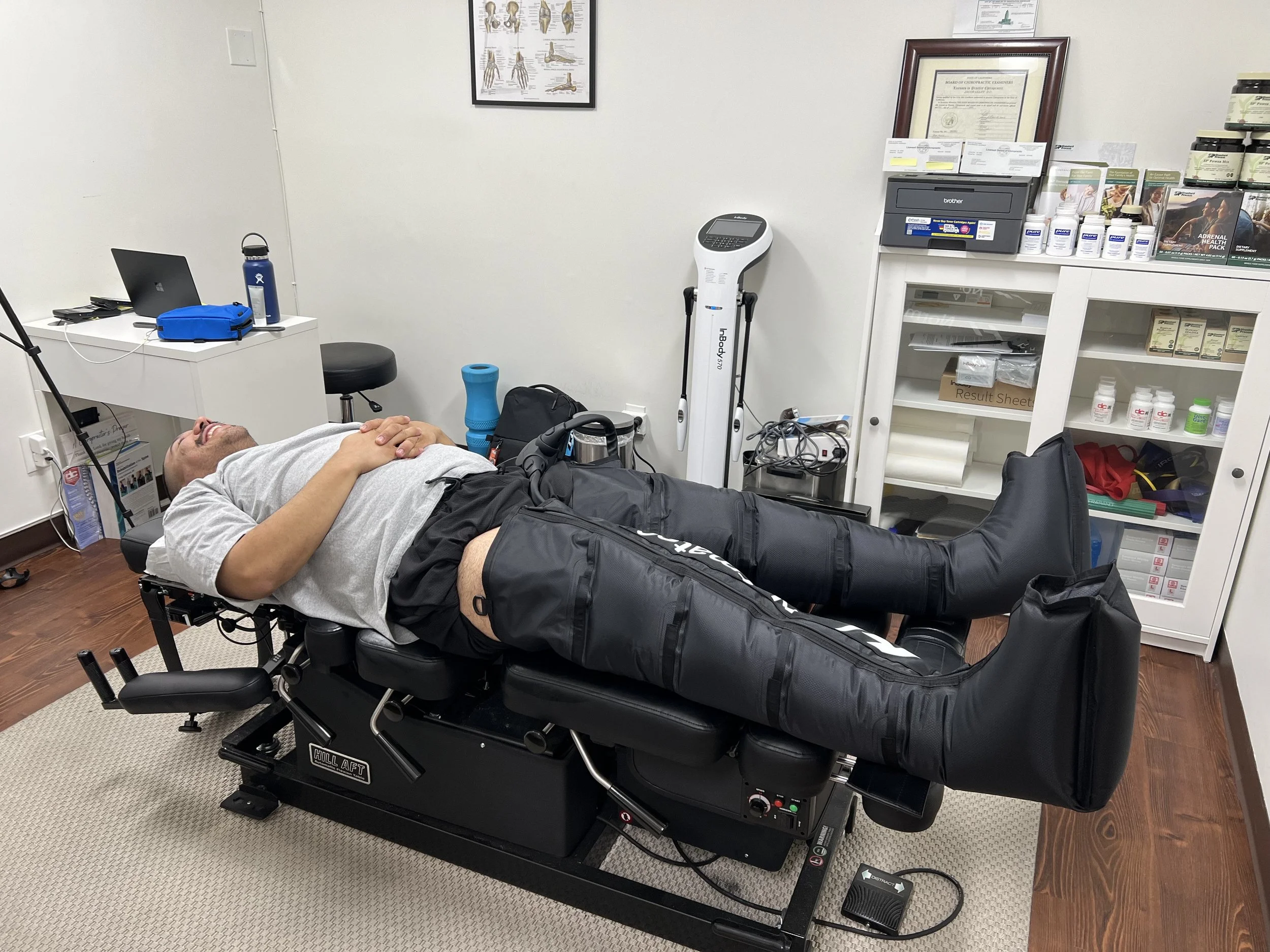 Man lying on a medical treatment table in a clinical office, smiling, wearing grey t-shirt and black shorts, with a leg injury or brace.