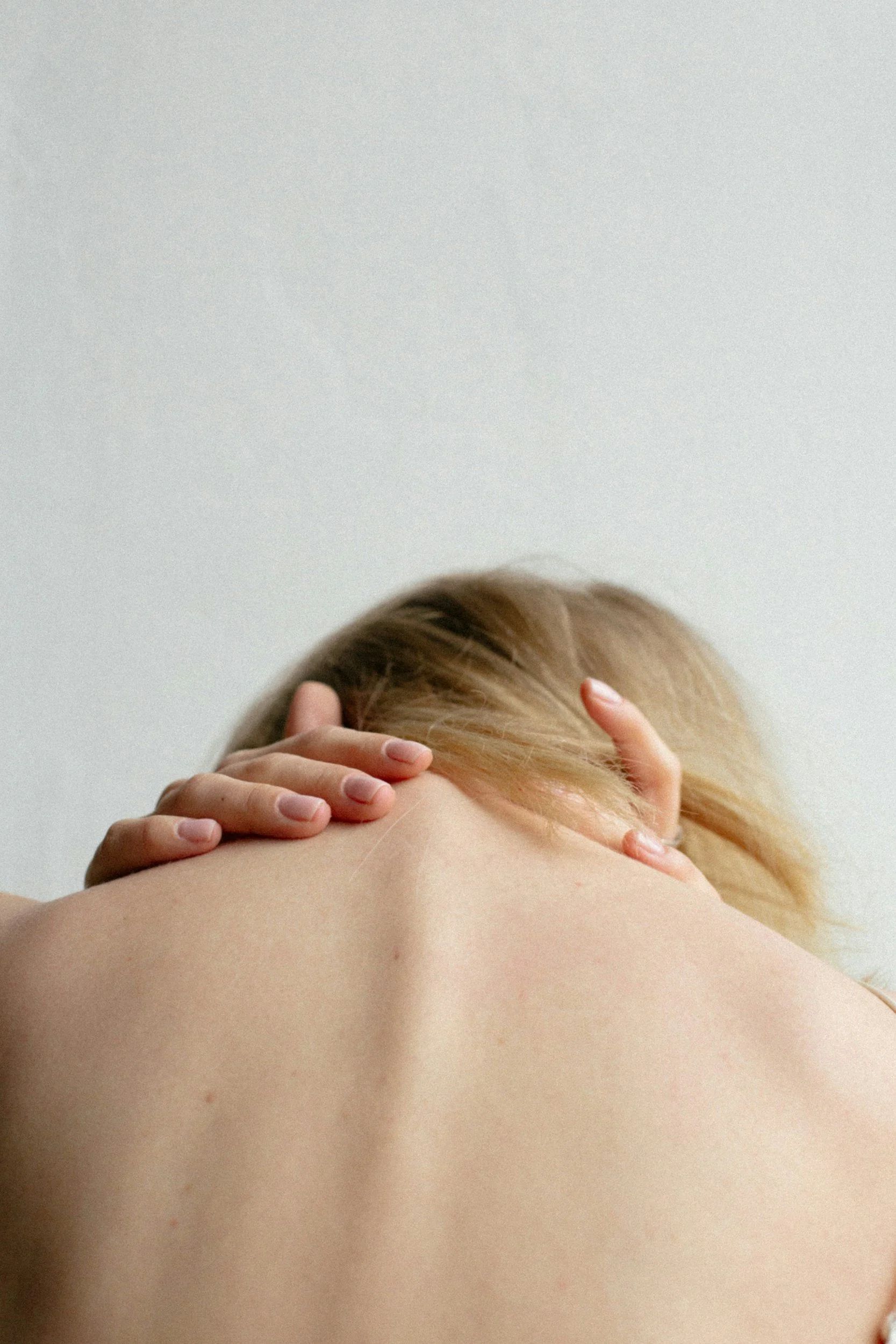 A person with light skin and blonde hair holding their head with both hands, seen from the back, with a plain white wall in the background.