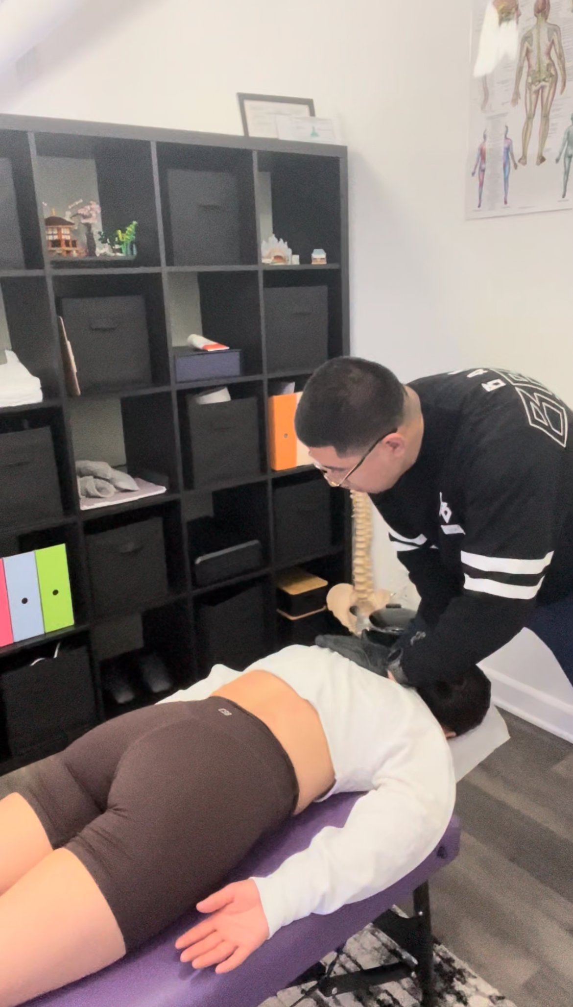 A chiropractor adjusting a woman's neck during a session in an office with bookshelf and anatomy poster.