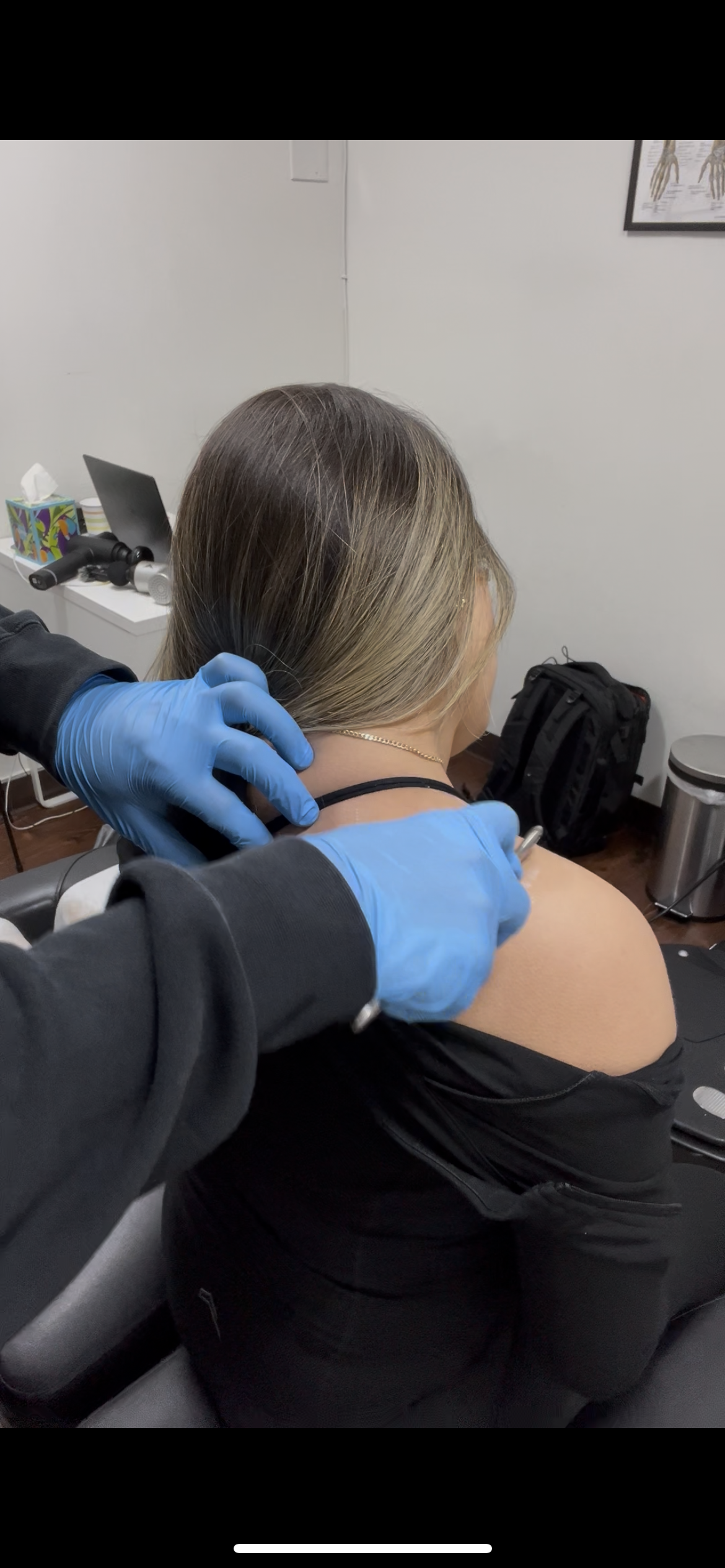A woman receiving soft tissue work on her shoulder from a healthcare professional wearing blue gloves, in an office or clinic setting.