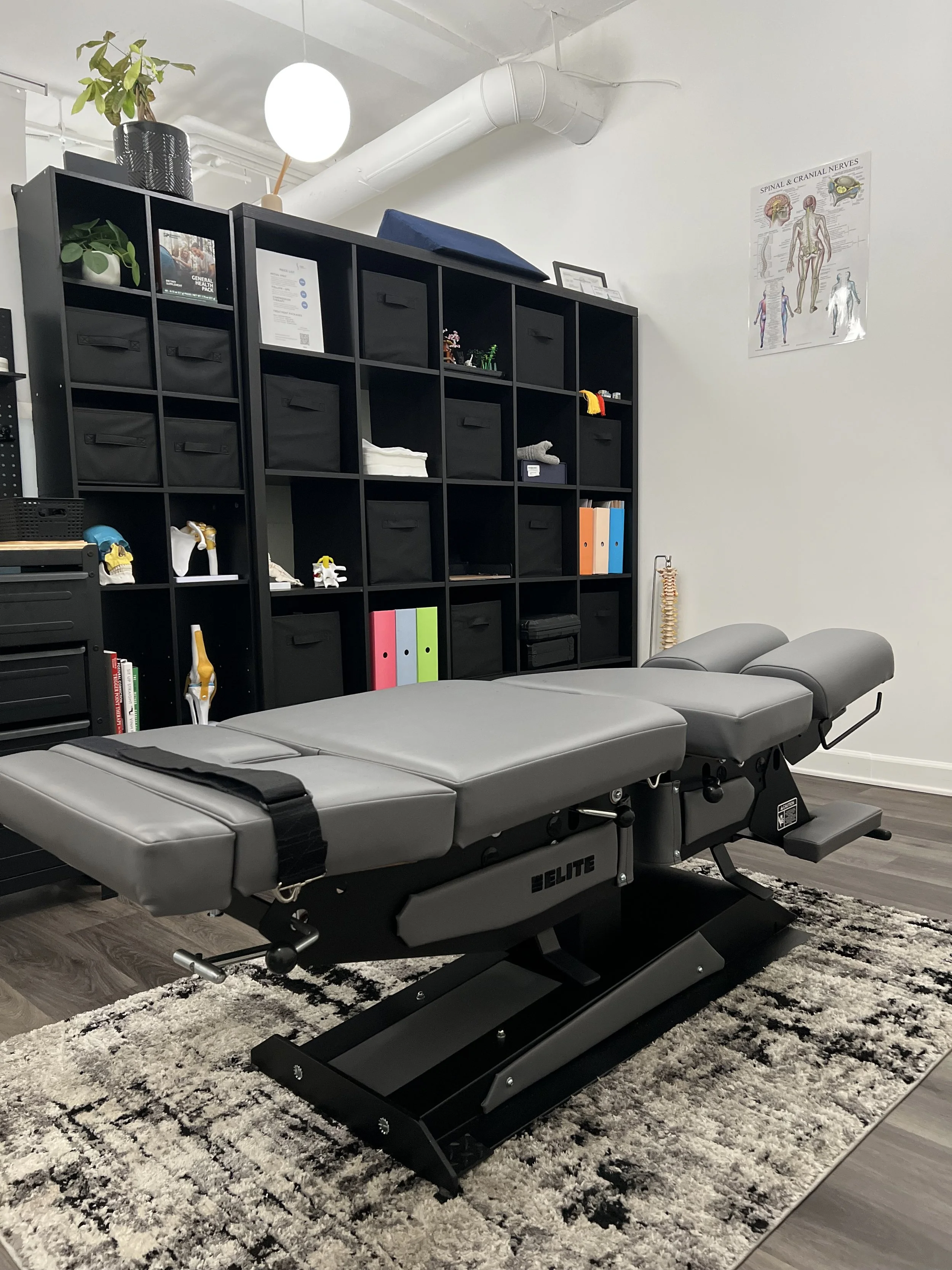 A spinal decompression table in a medical office with a black cubed shelving unit against a white wall, a poster showing human nervous system on the wall, and a gray rug on a wood floor. Located in encino california.