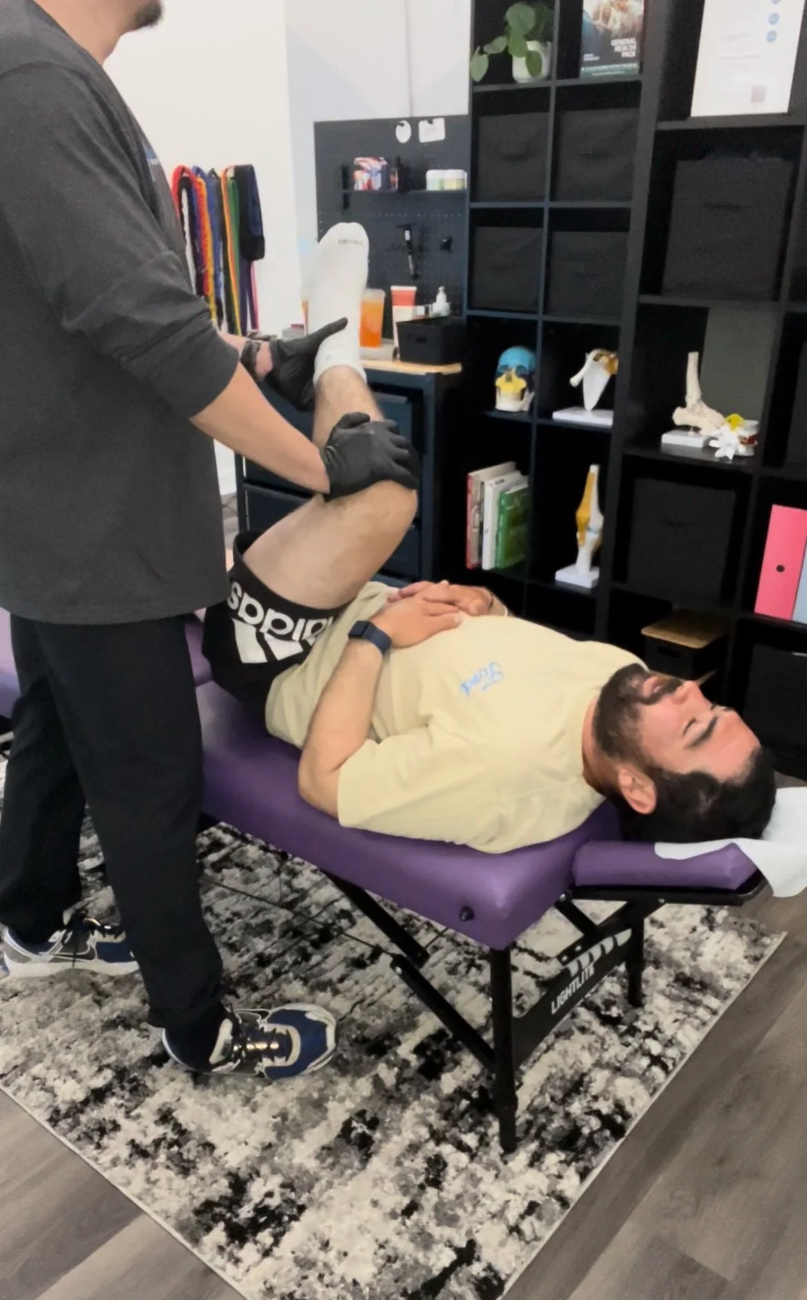 A man receiving physical therapy or chiropractic treatment lying on a treatment table while a therapist holds his leg. The room has black shelving with medical models, books, and supplies.