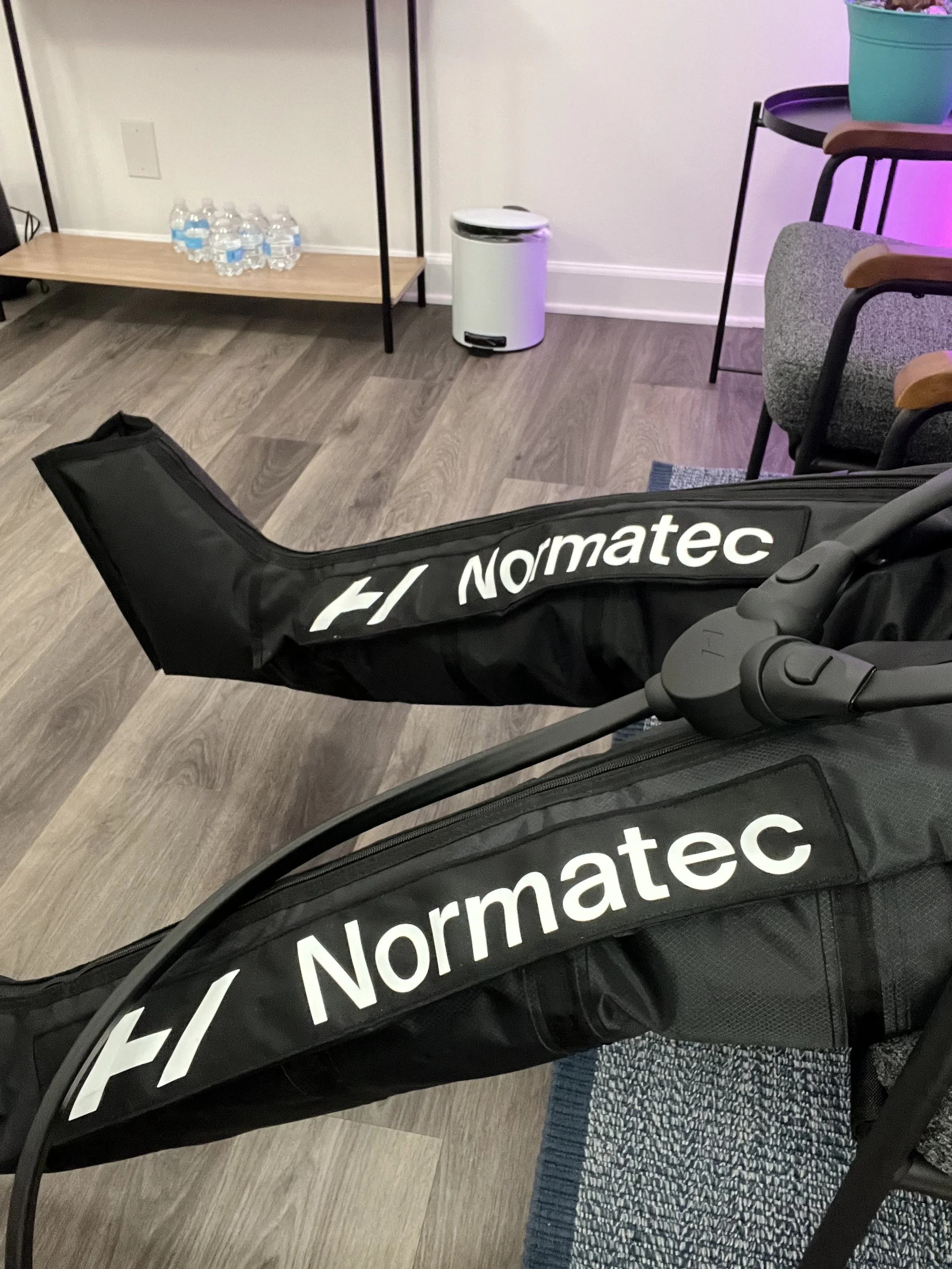 Indoor room with two Normatec compression therapy sleeves, a small wooden table with water bottles, a trash can, chairs, and a teal flower pot with pink flowers.