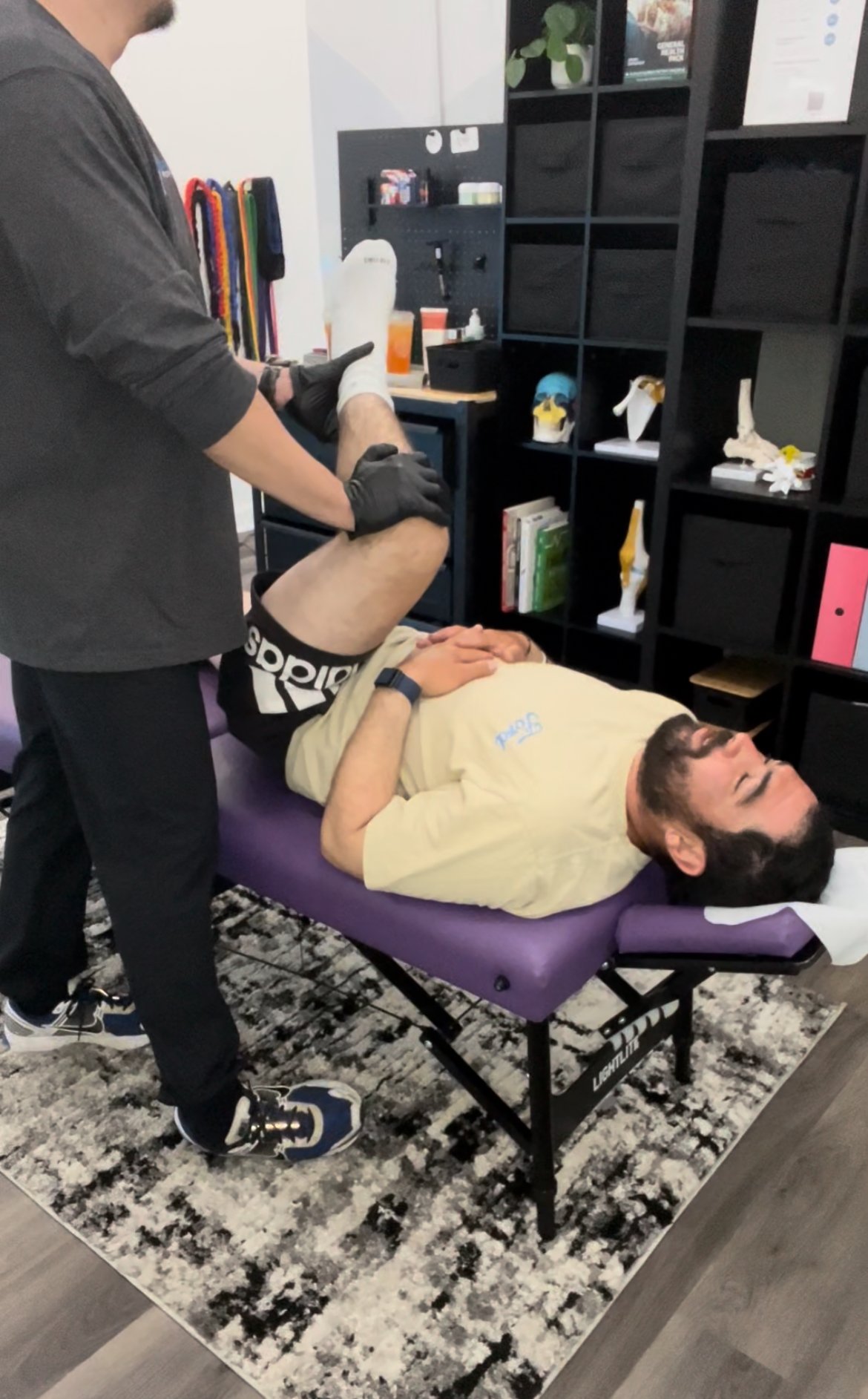 A man lying on a massage table receiving physical therapy or chiropractic treatment from a practitioner in a room with black shelving and books.